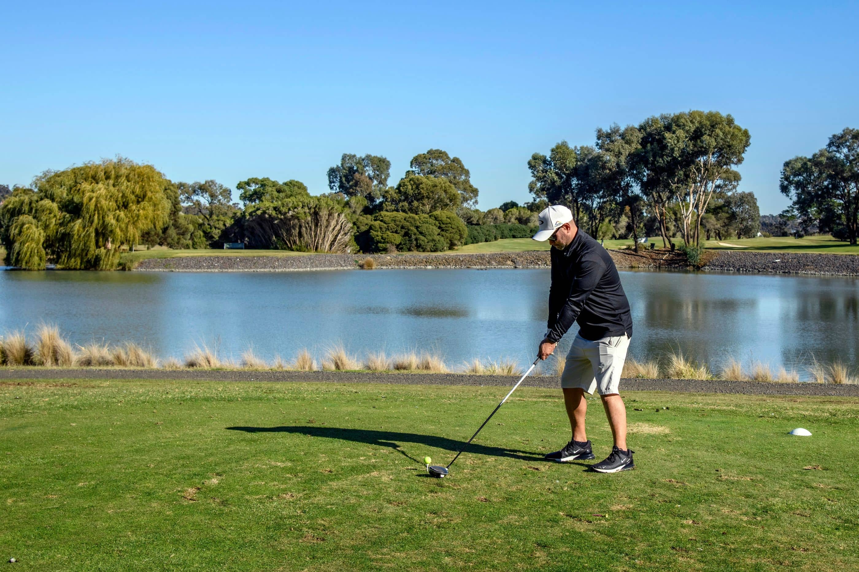 Man with golf club on tee box by a lake, with trees in the background