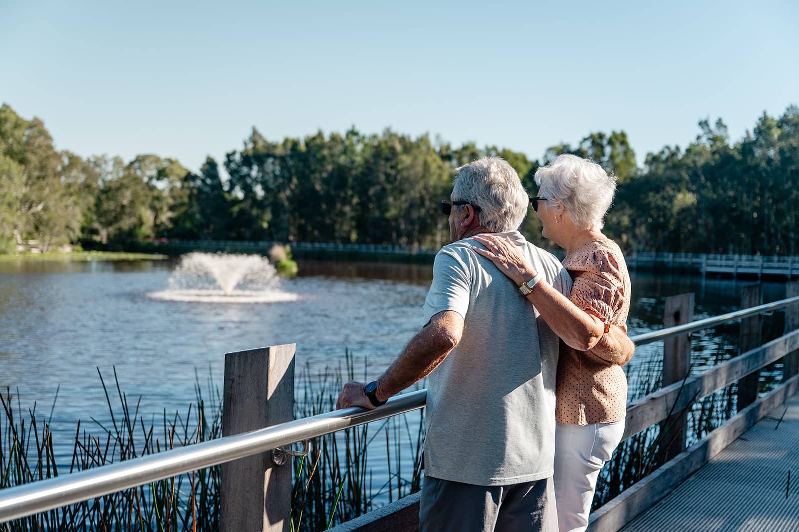 Couple standing on a boardwalk overlooking a lake with a fountain, in an Ingenia Lifestyle community.
