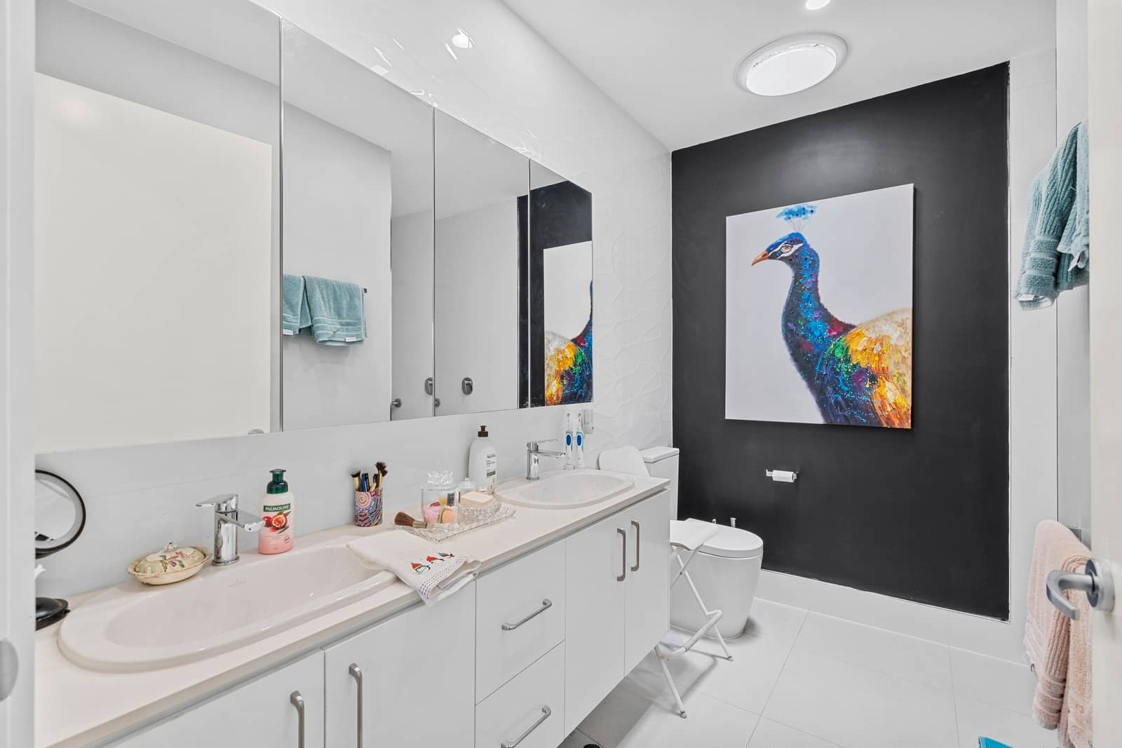 A modern bathroom with a double vanity, a peacock painting on a black accent wall, and white tiled floors.