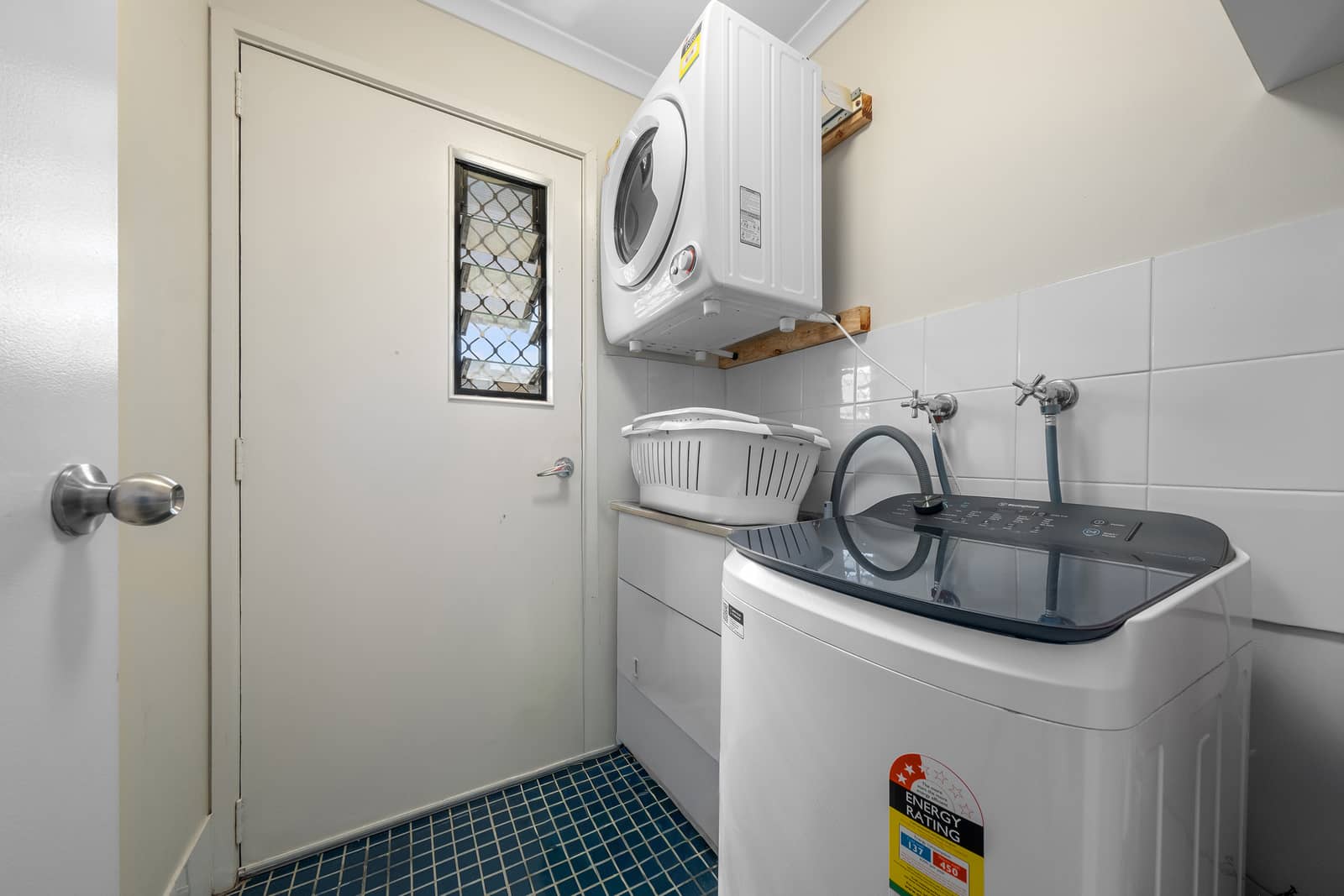 A top-load washing machine, laundry basket, and wall-mounted dryer in a small laundry room.