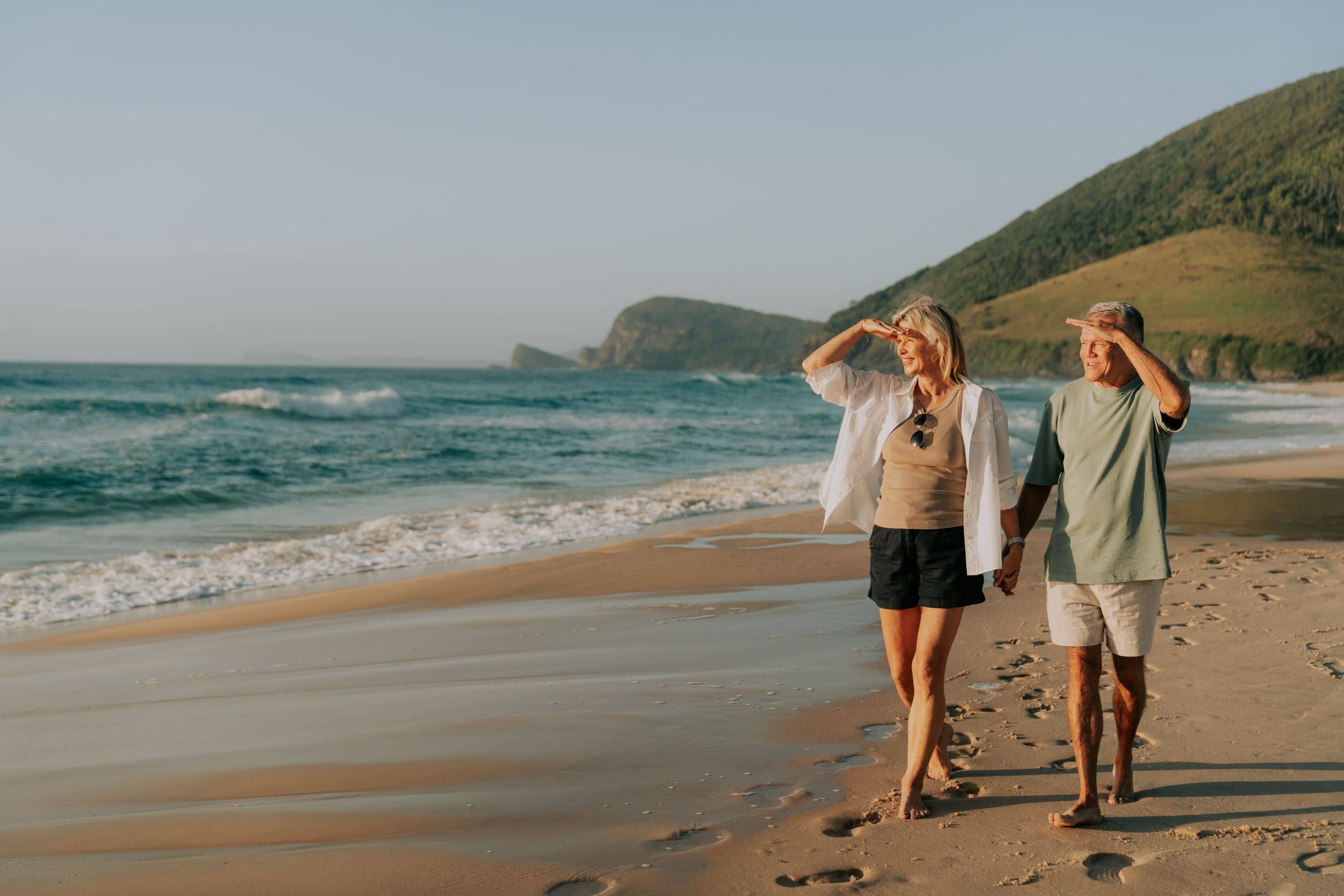 Two people walk barefoot on a sunlit beach with waves, shading their eyes while looking at the distant hills.