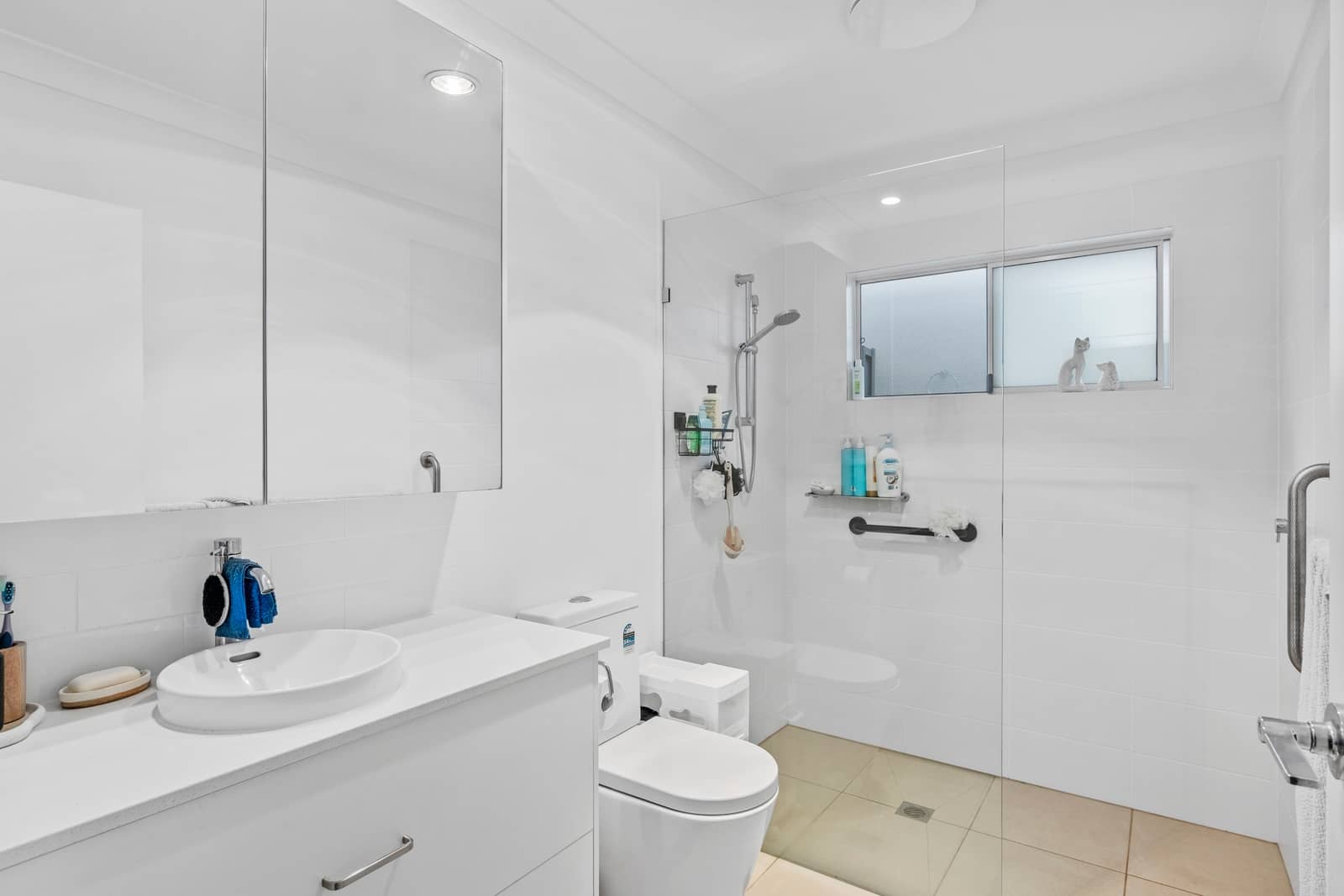 White tiled bathroom featuring a vanity, toilet, and walk-in shower with a glass screen and grab bars, suitable for over-55 living.