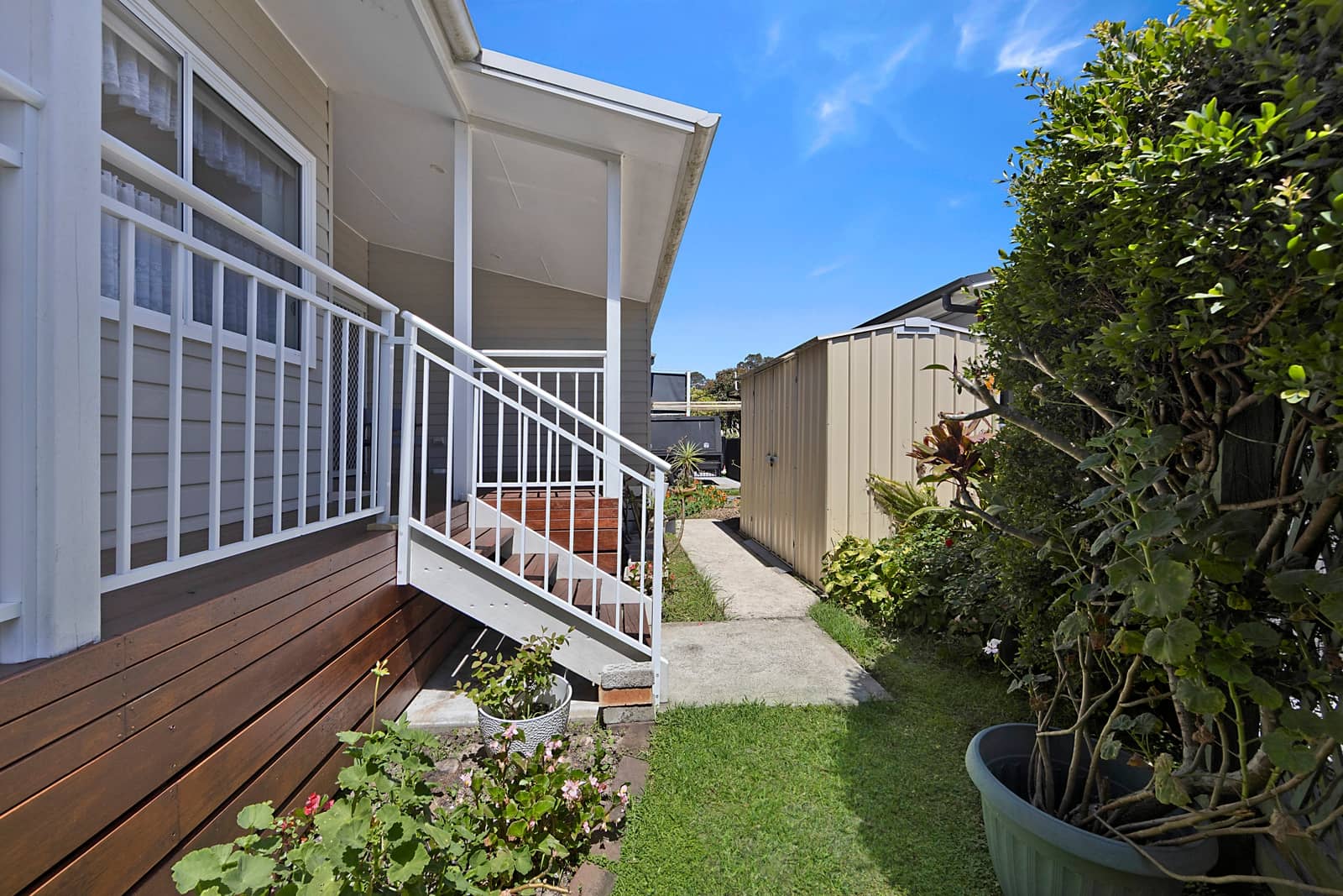 Exterior view of an Ingenia Lifestyle home featuring a covered timber deck, white railings, a small garden, and a detached storage shed.
