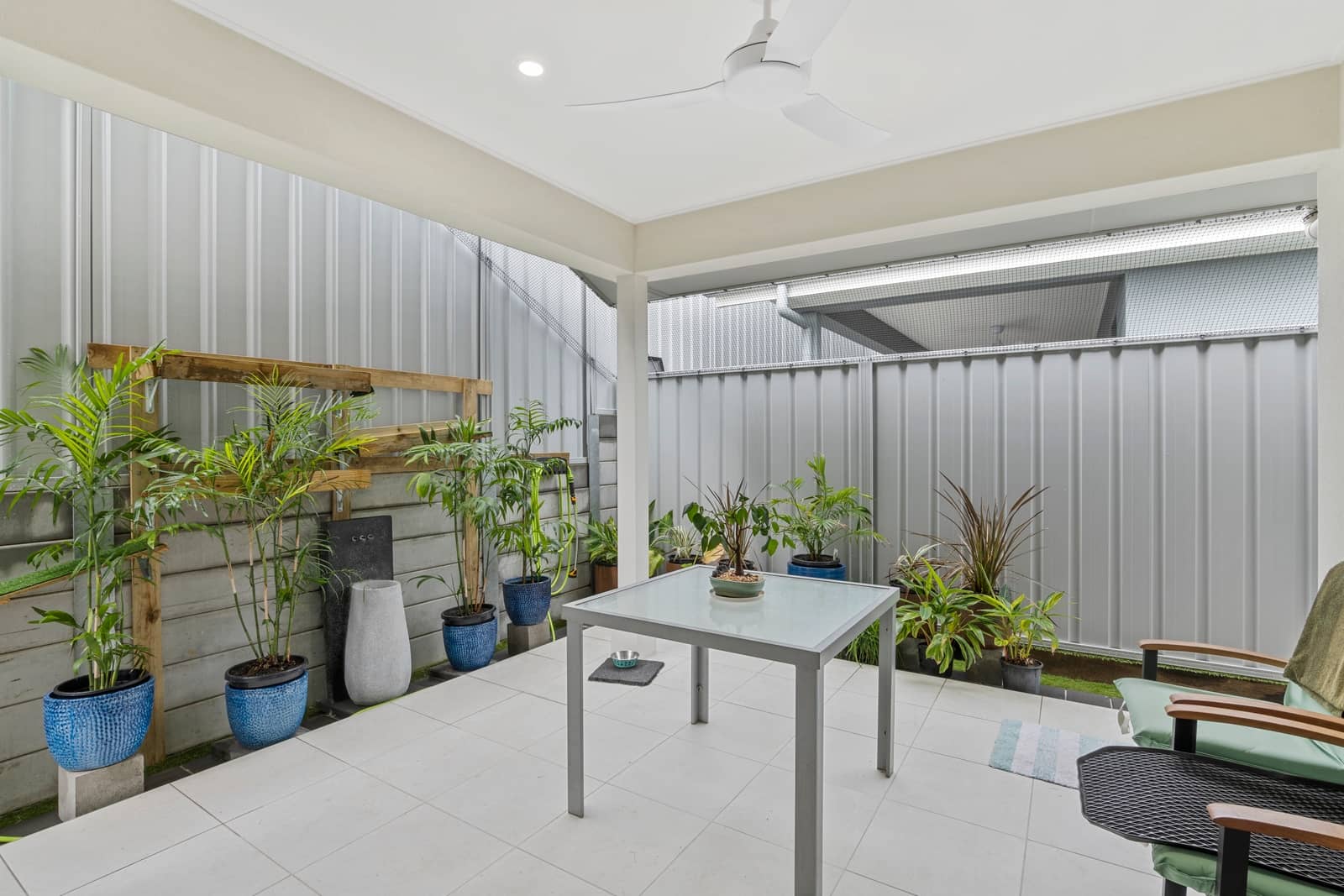 Covered patio with a table, chairs, and numerous potted plants against a metal fence, offering low-maintenance living.