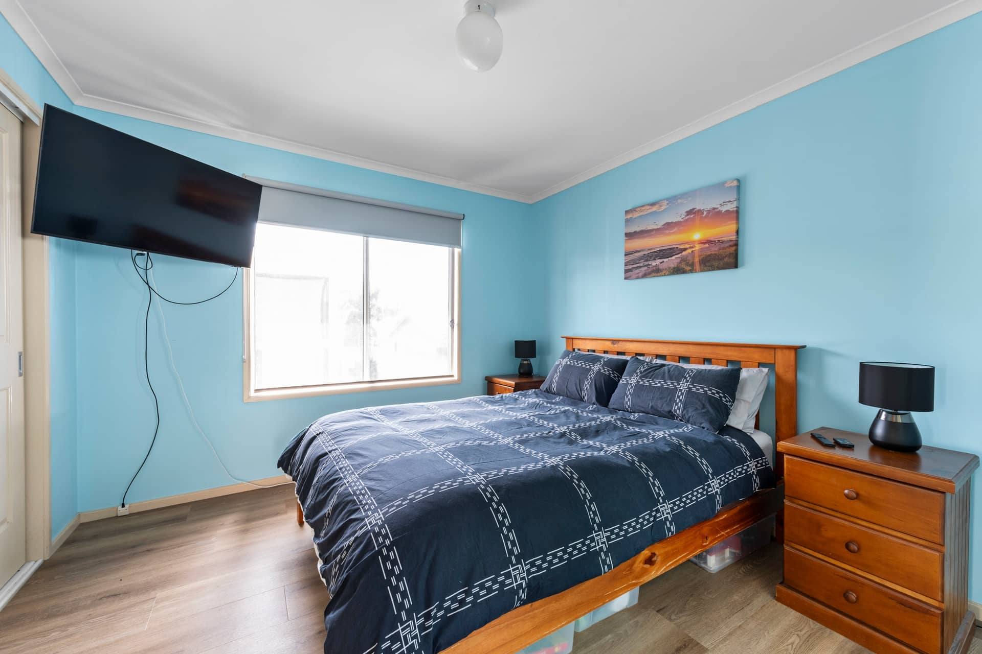 Bedroom with light blue walls, wood-look flooring, a wooden bed with dark blue bedding, bedside tables, and wall-mounted TV.