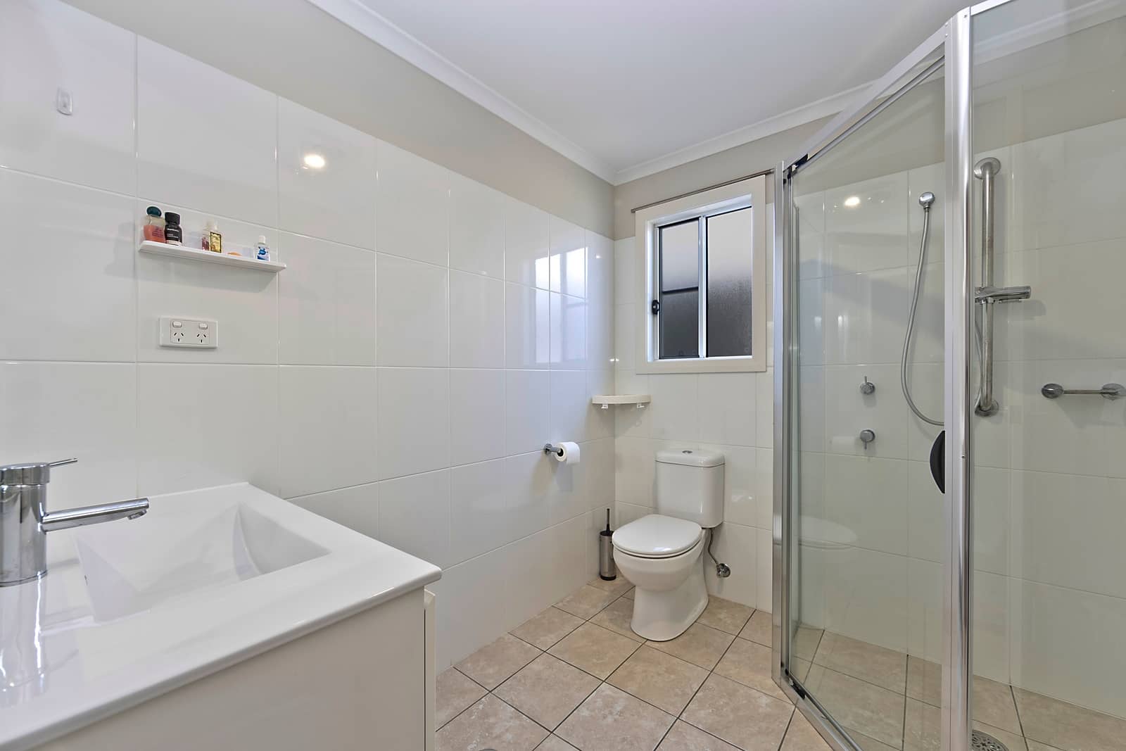 Modern bathroom with white wall tiles, beige floor, vanity, toilet, and glass shower with grab bar in an Ingenia Lifestyle home.