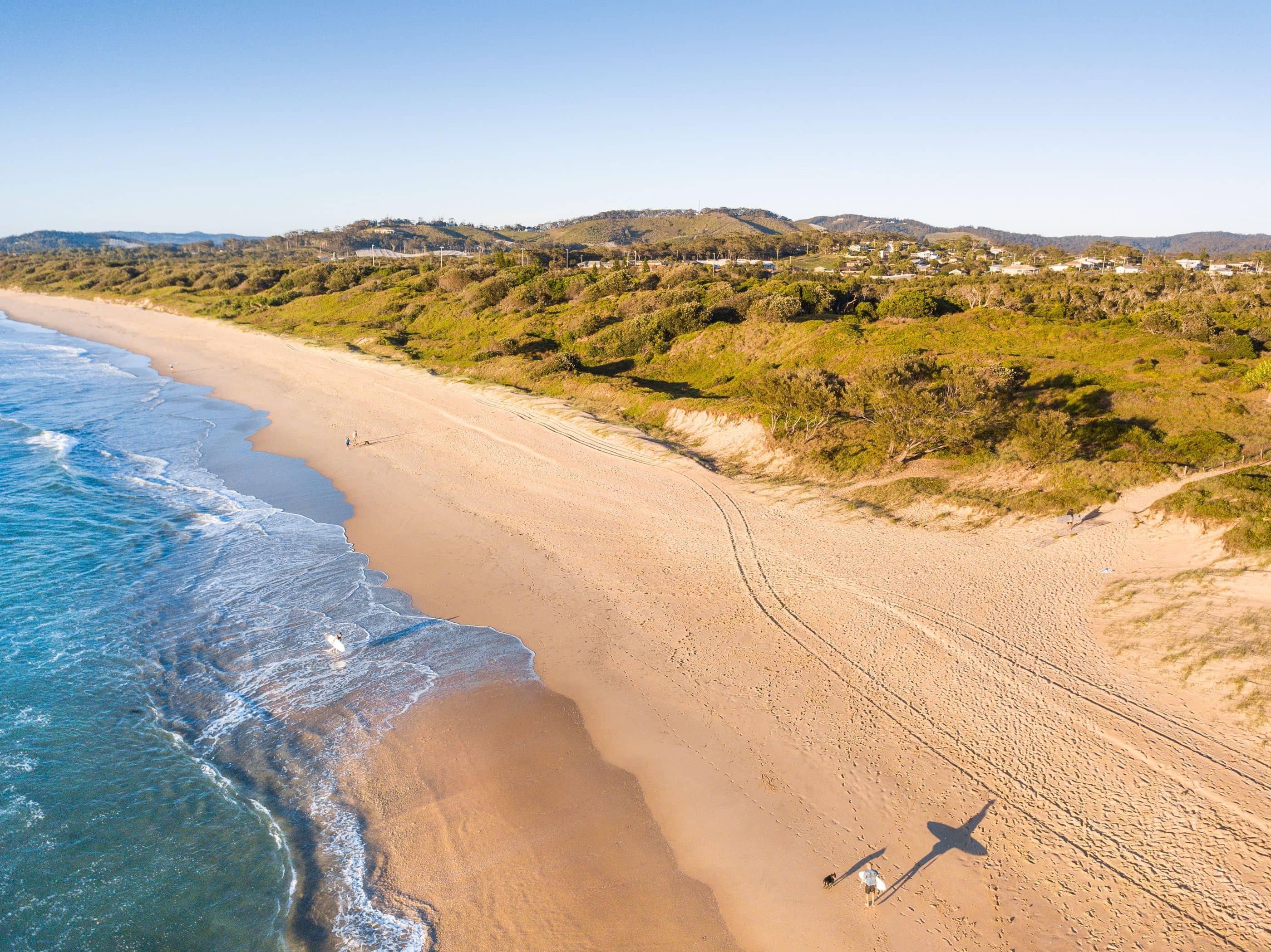 Coastal beach with rolling hills and houses in the background and people walking on the sand.