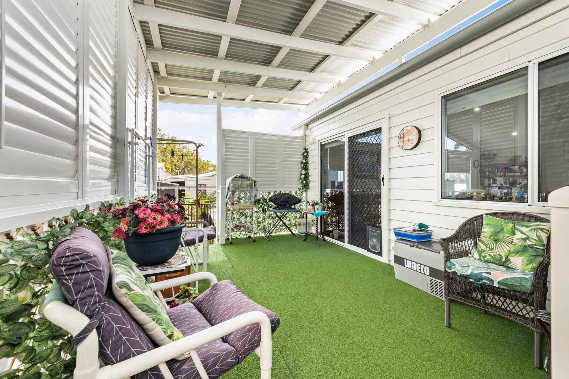 Covered patio with artificial grass, louvered shutters, outdoor seating, and a BBQ, part of an Ingenia Lifestyle home.