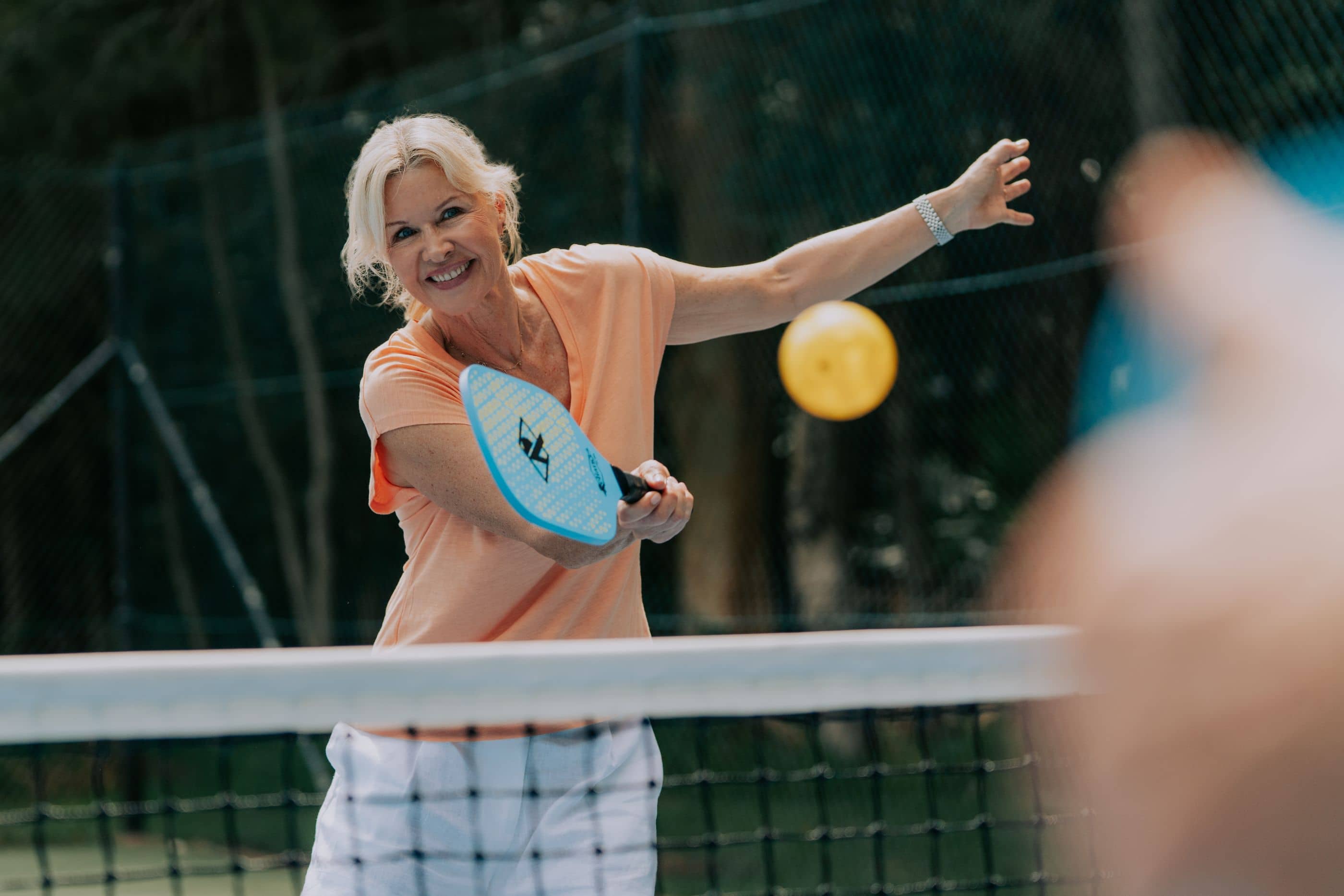 A smiling woman playing pickleball with a blue paddle and yellow ball on a court at an Ingenia Lifestyle community.