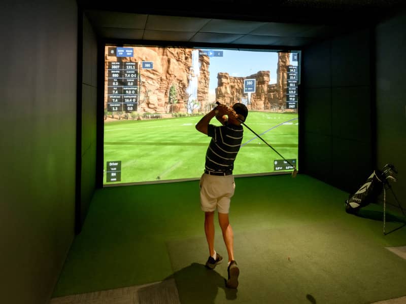 Person swings a golf club in a simulator room with a digital golf course displayed on a large screen.