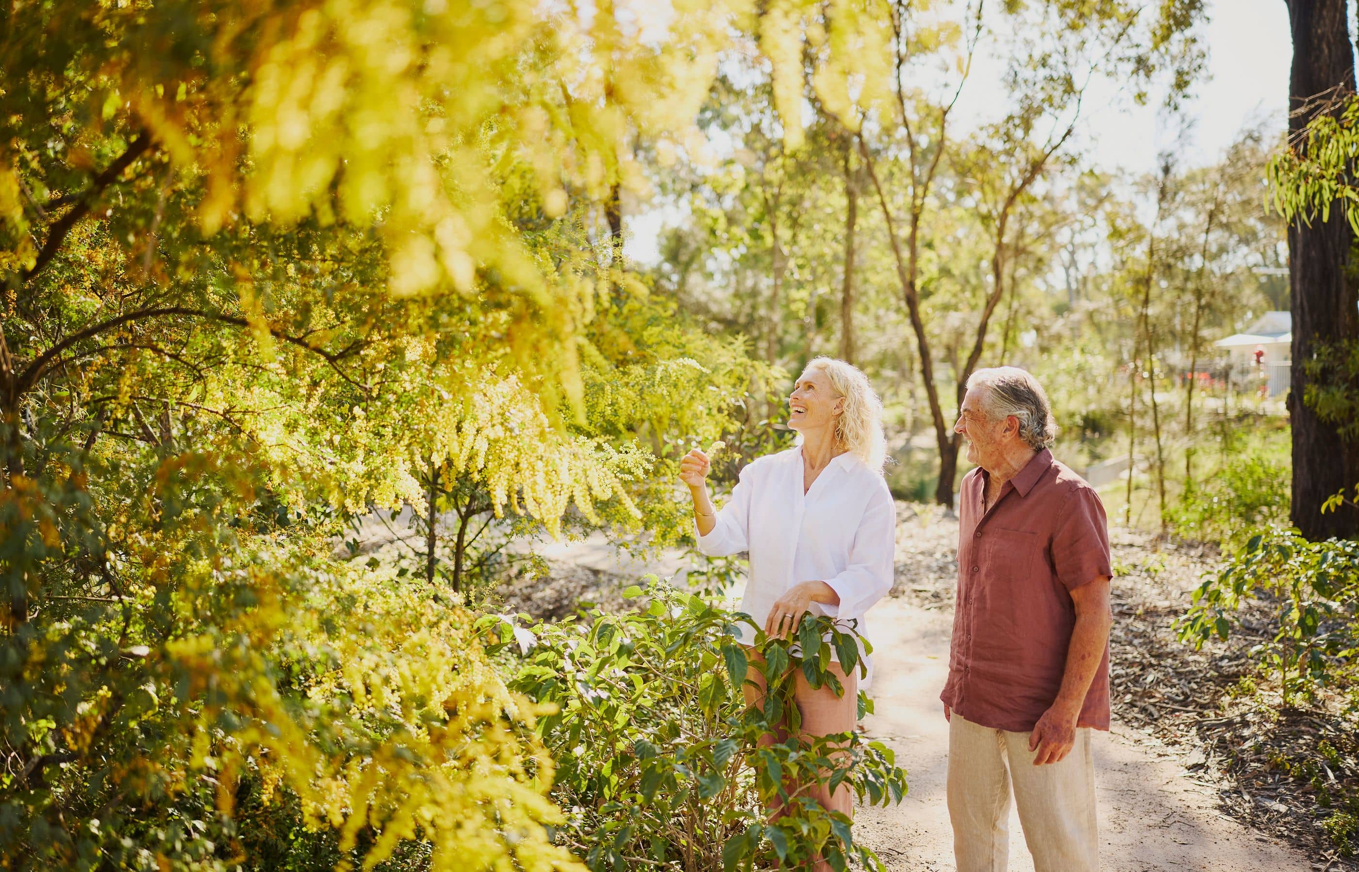 A woman and a man stand amidst lush greenery and flowering trees in an Ingenia Lifestyle community.