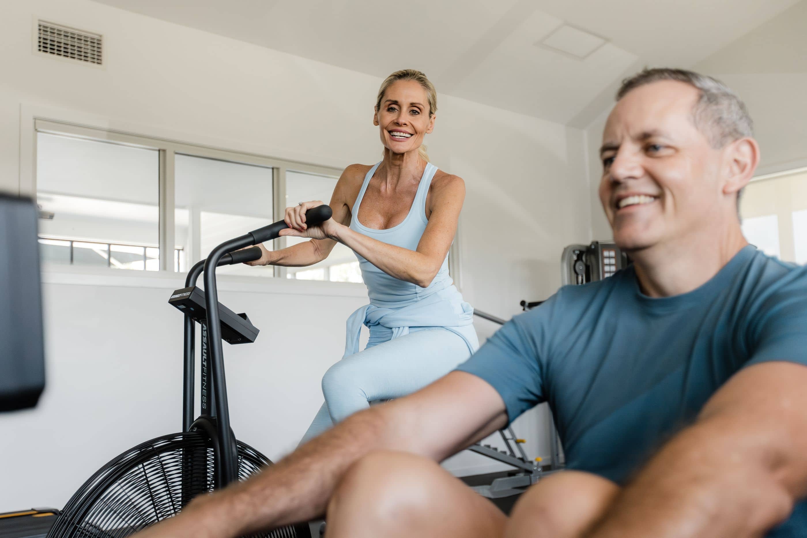 A woman on an air bike smiles at the camera, with a man in the foreground.