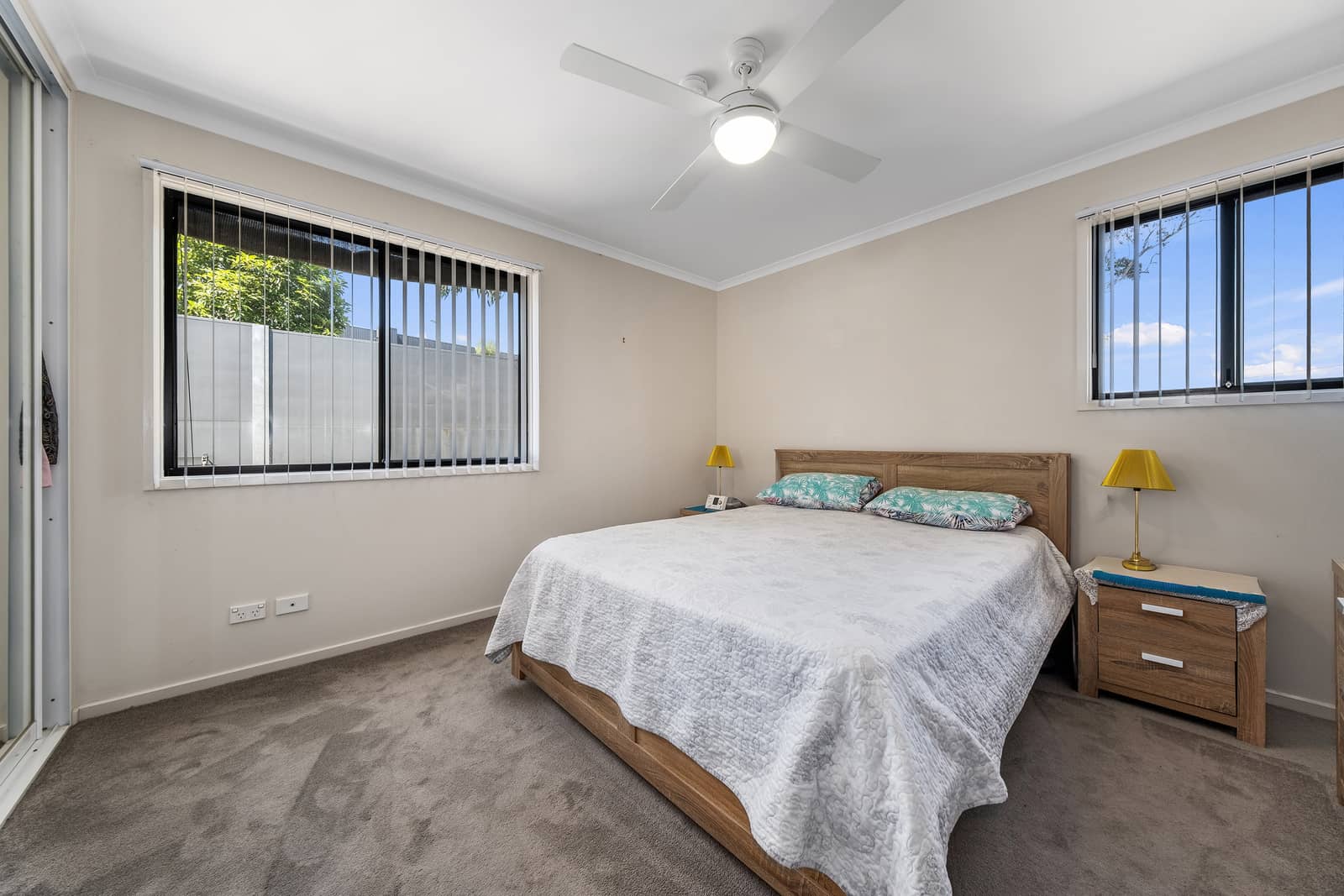 A bedroom in an Ingenia Lifestyle community featuring a queen bed, a ceiling fan, and two windows with blinds.