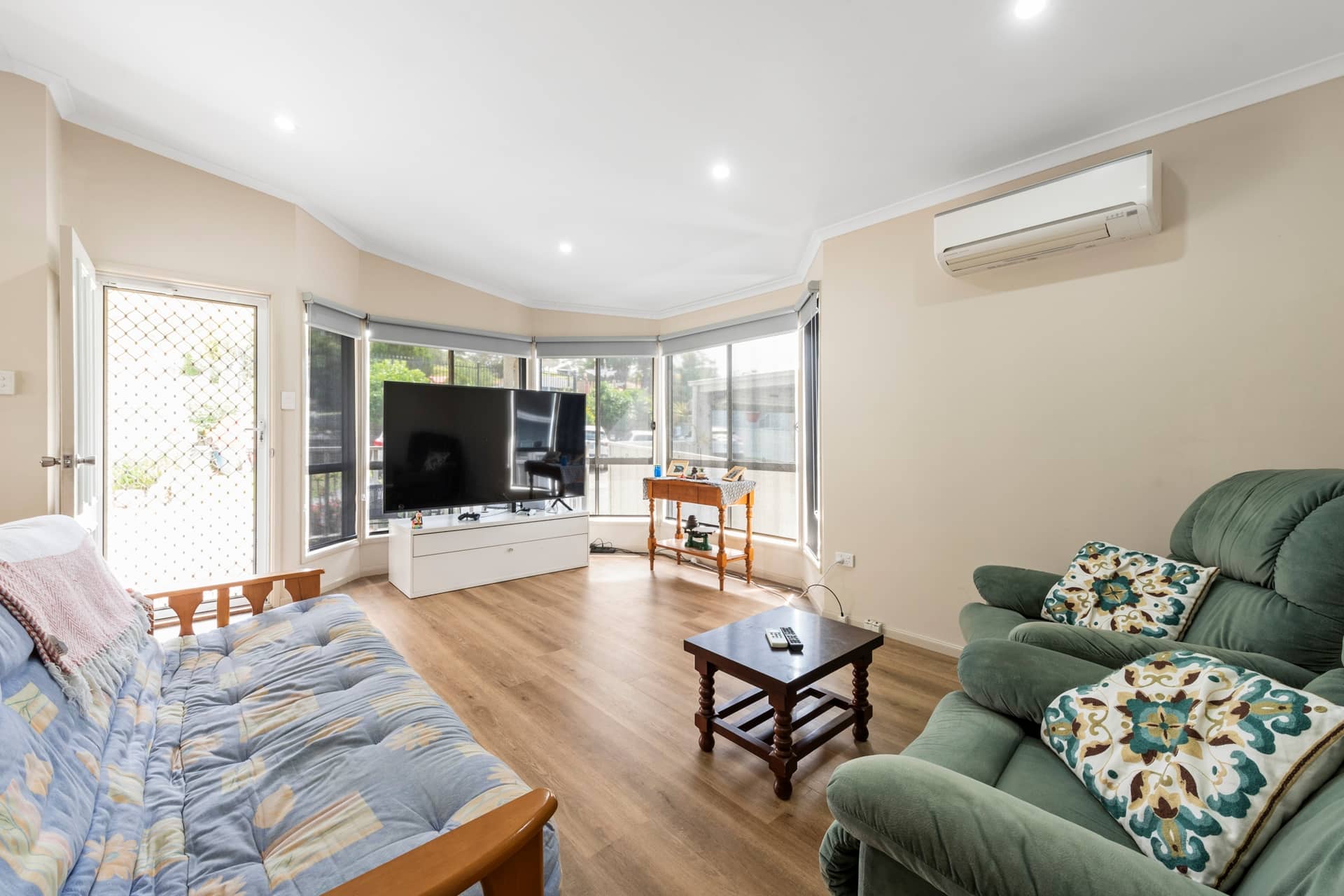 Spacious living room with wood-look flooring, large bay window, TV, green armchairs, and a daybed in an Ingenia Lifestyle home.