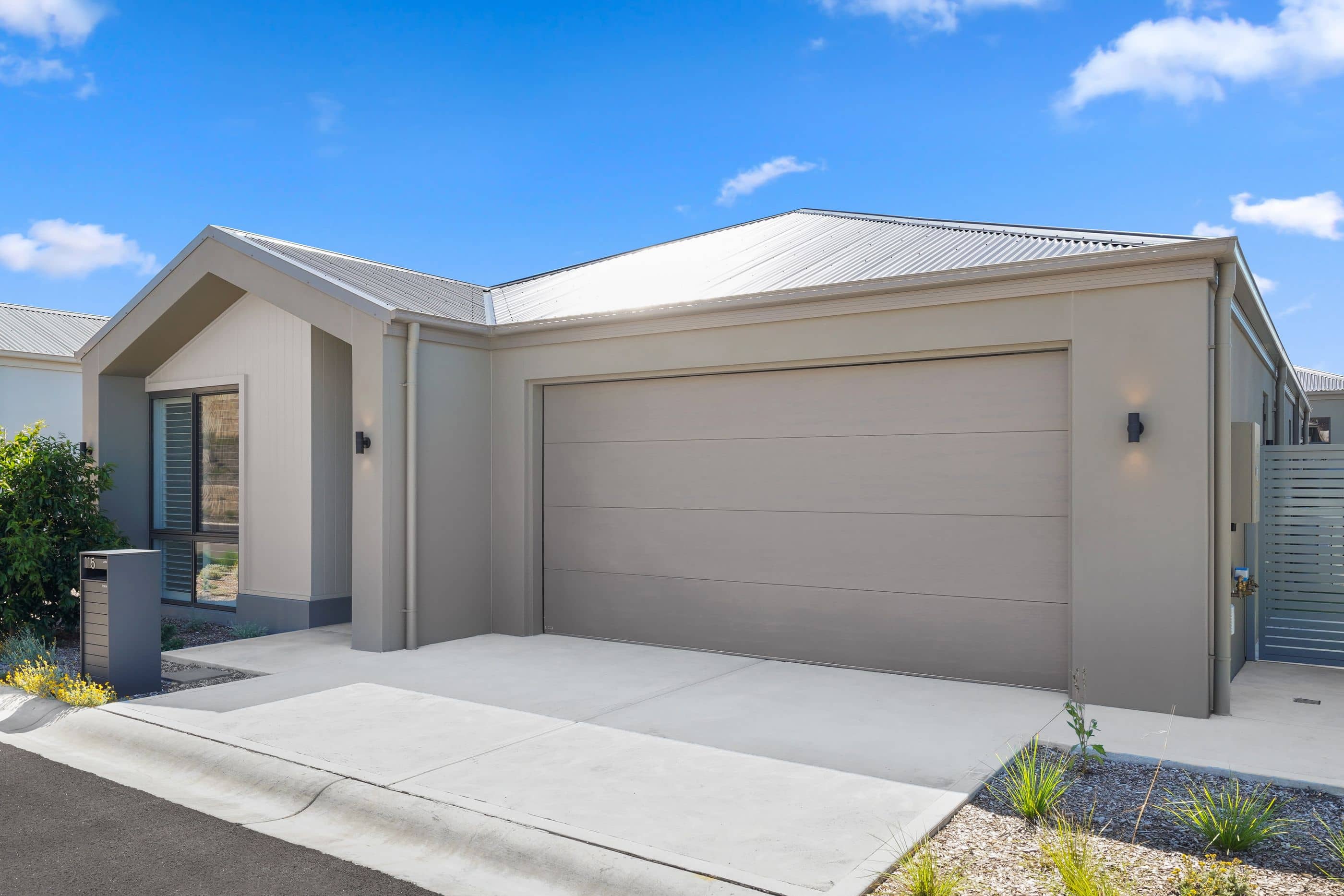 A contemporary Ingenia Lifestyle home with a grey garage, landscaped garden, and concrete driveway under a blue sky.