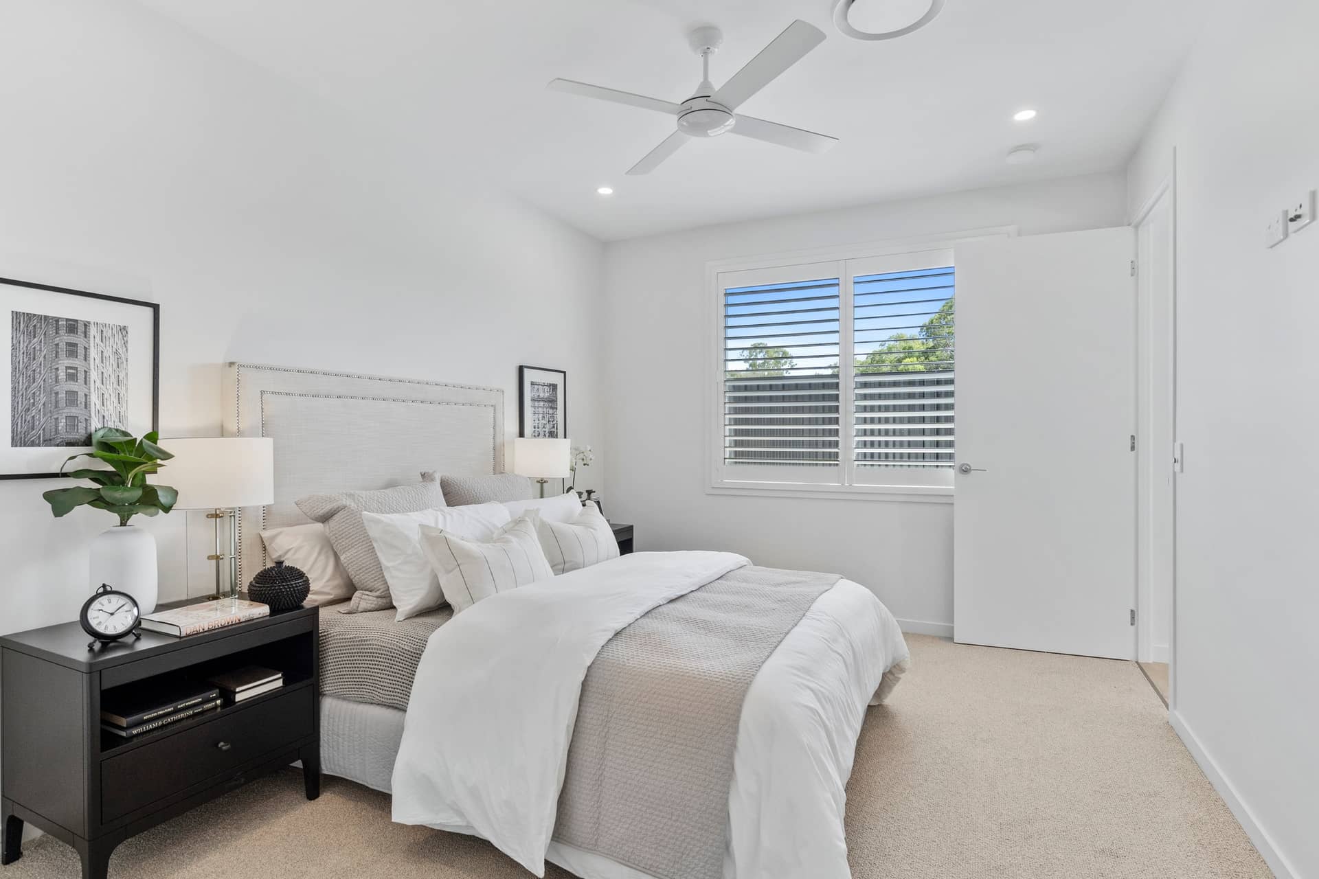 A bright bedroom with a white bed and grey accents, featuring a bedside table with a plant and a framed picture.