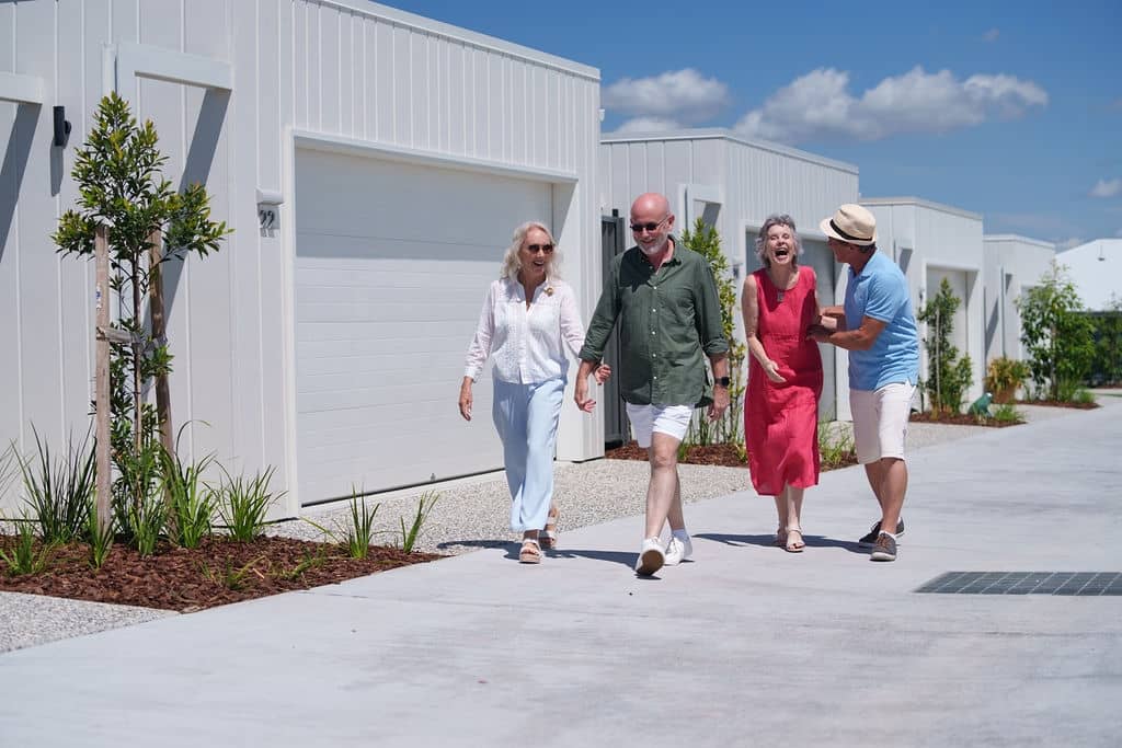 Four people laughing while walking on a pathway past white, low-maintenance homes in an Ingenia Lifestyle community.