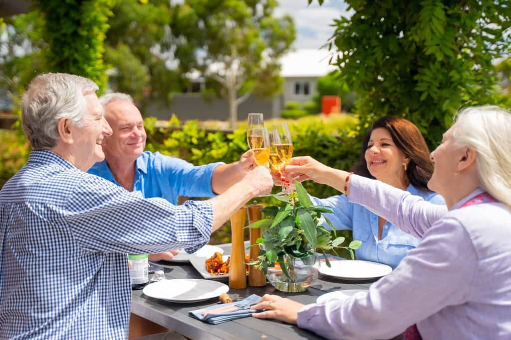 Four smiling individuals toasting with champagne flutes at an outdoor table in an Ingenia Lifestyle community.