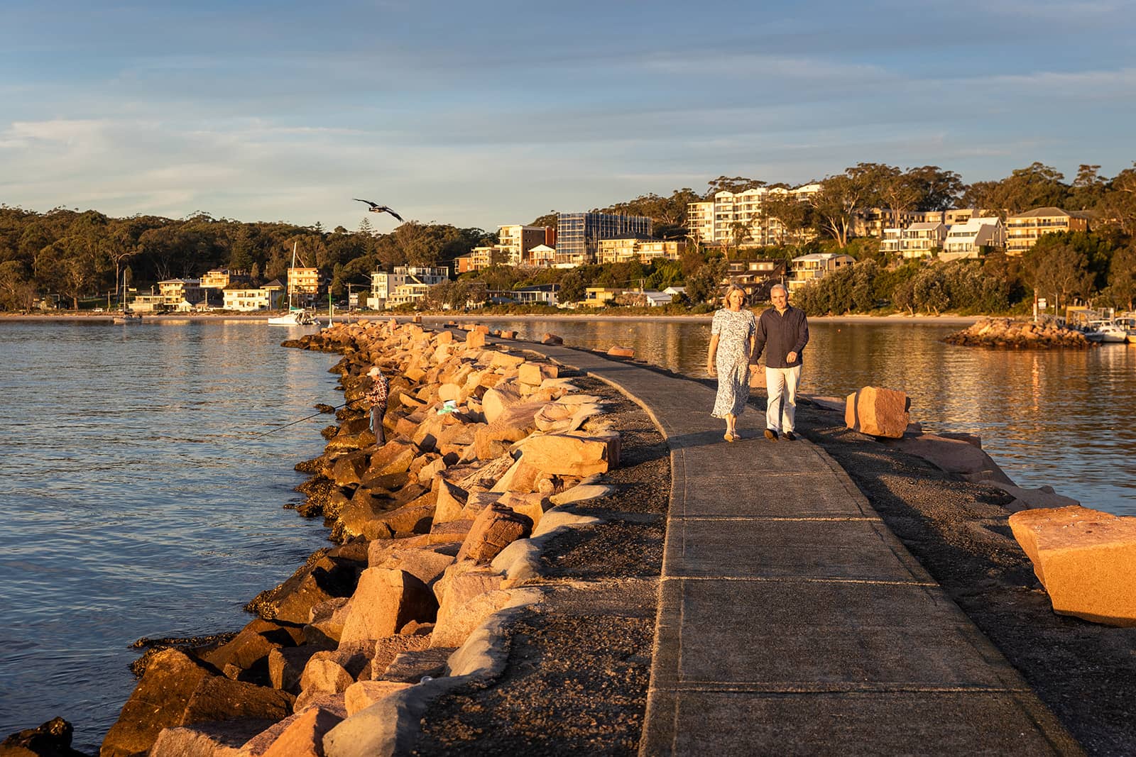 A couple walks on a coastal breakwater path at sunset, with a person fishing from the rocks and a town along the bay.