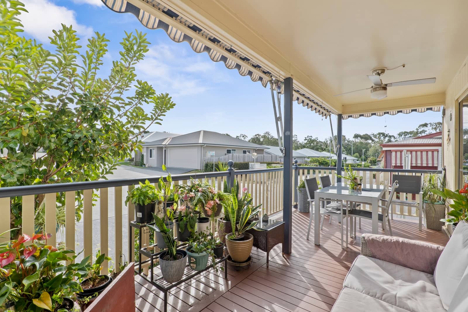 Covered outdoor deck with dining set, couch, and potted plants, overlooking other homes in an Ingenia Lifestyle community.