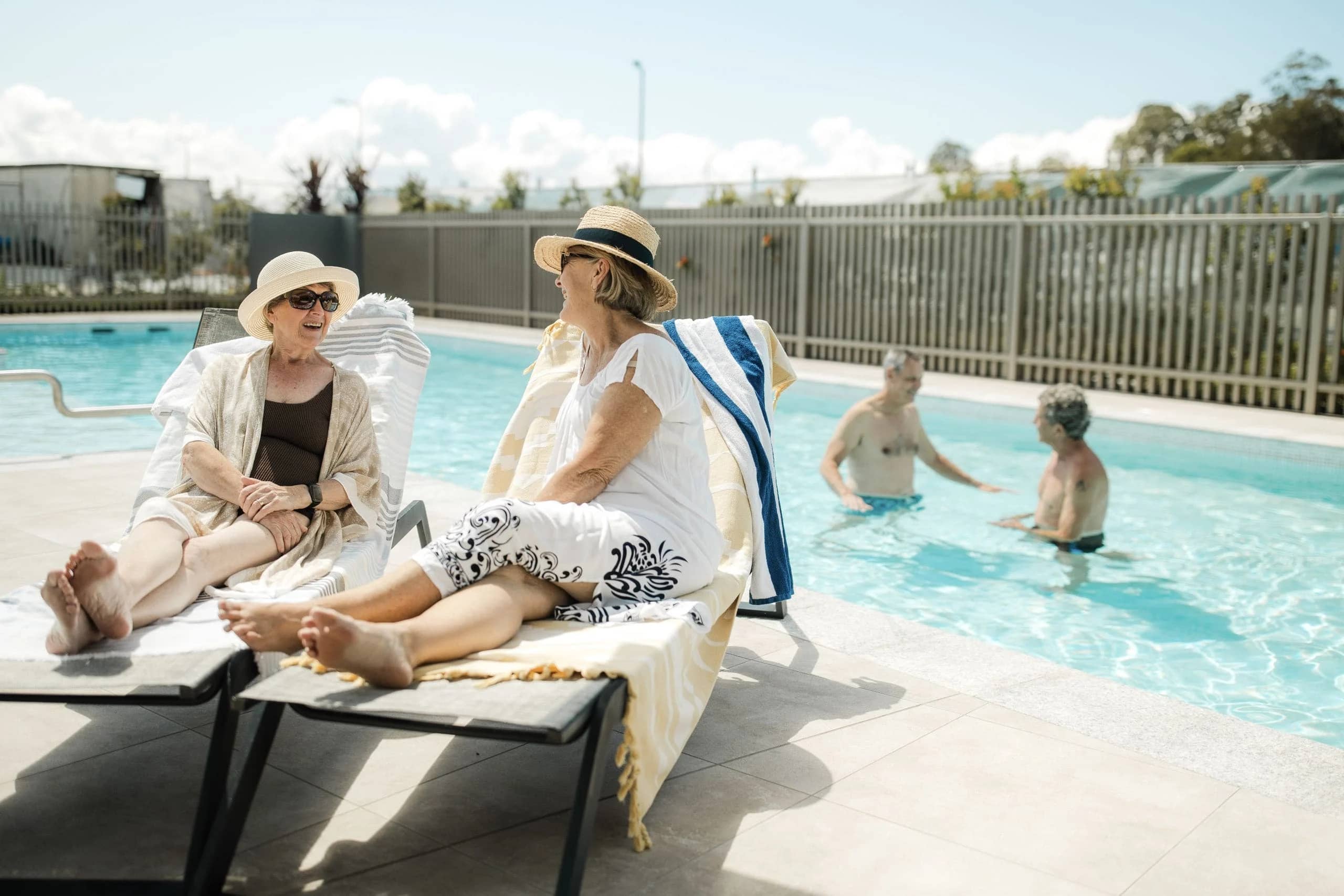 Two women relax in sun loungers by a pool, while two men swim in the background at an Ingenia Lifestyle community.
