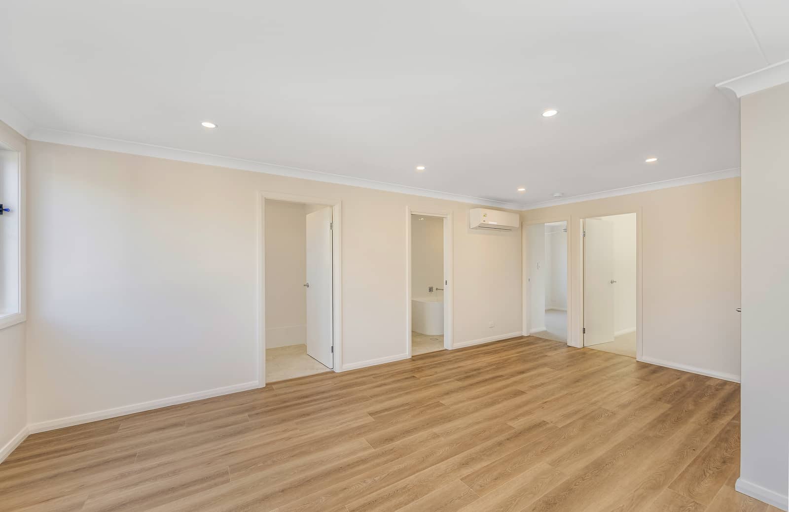 Empty room in an Ingenia Lifestyle home, featuring wood-look flooring, light walls, recessed lights, an air conditioner, and open doorways to other spaces.