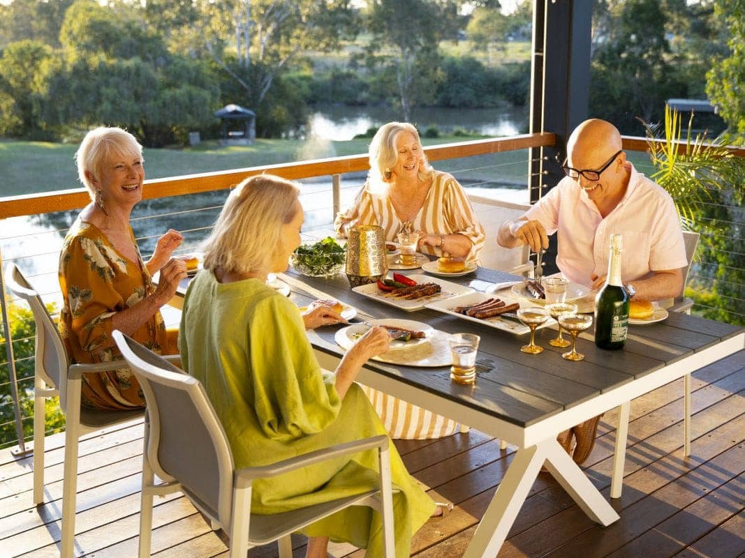 Four people enjoy a meal at a table on a deck overlooking a scenic waterway and trees.