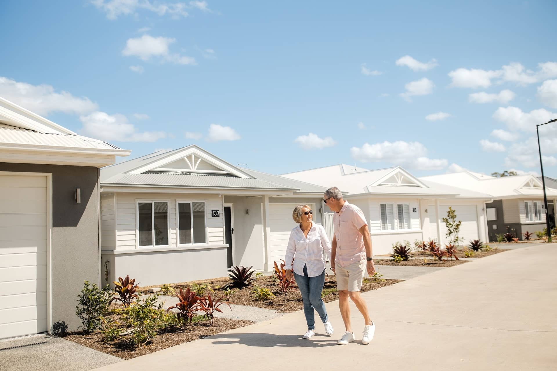 Two people walk on a path past modern land-lease homes in an Ingenia Lifestyle community, with landscaped gardens.