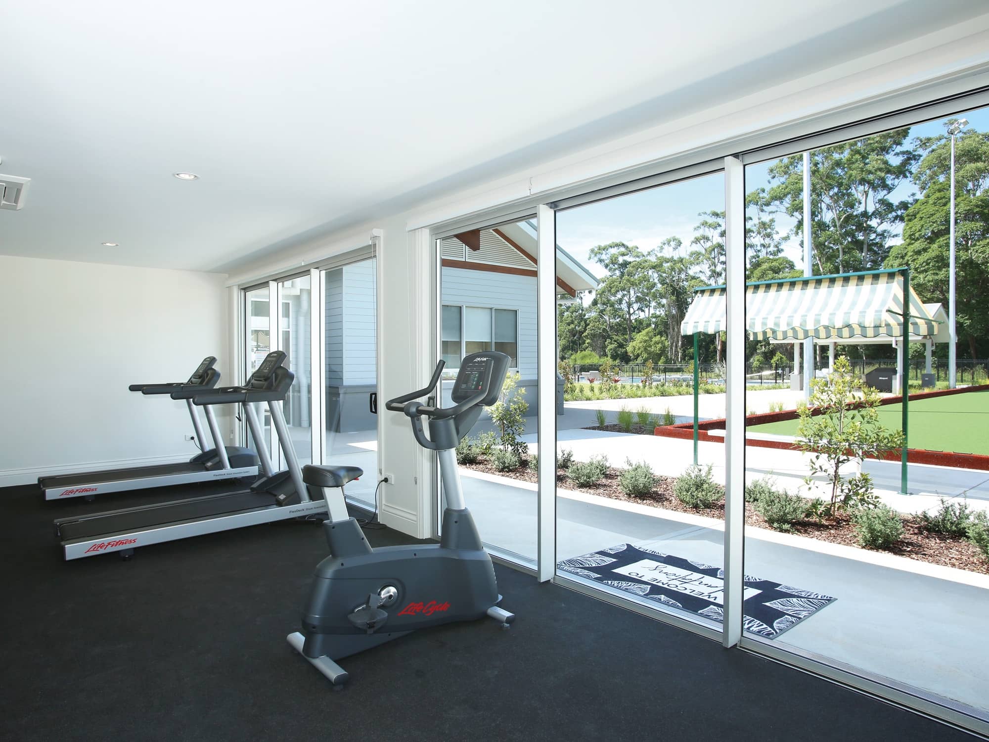 Two treadmills and an exercise bike in a gym with large sliding glass doors opening to community grounds.