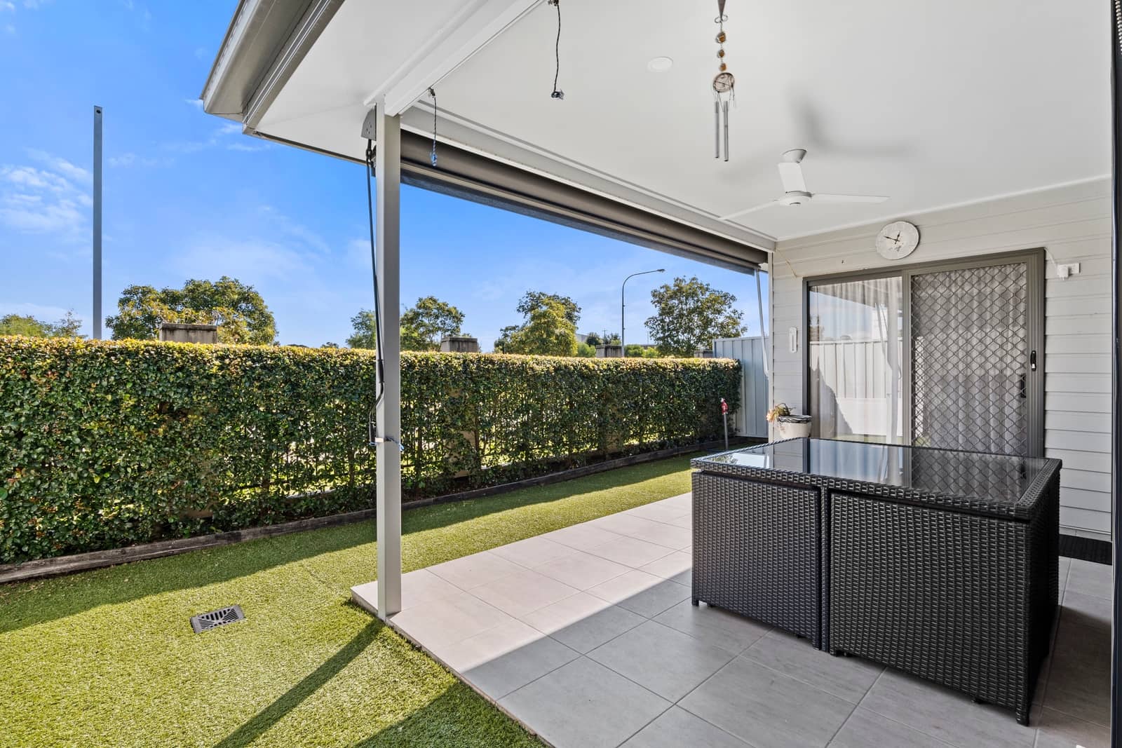 Covered outdoor tiled patio with a dining table and ceiling fan, leading to a low-maintenance lawn and hedge at an Ingenia Lifestyle home.