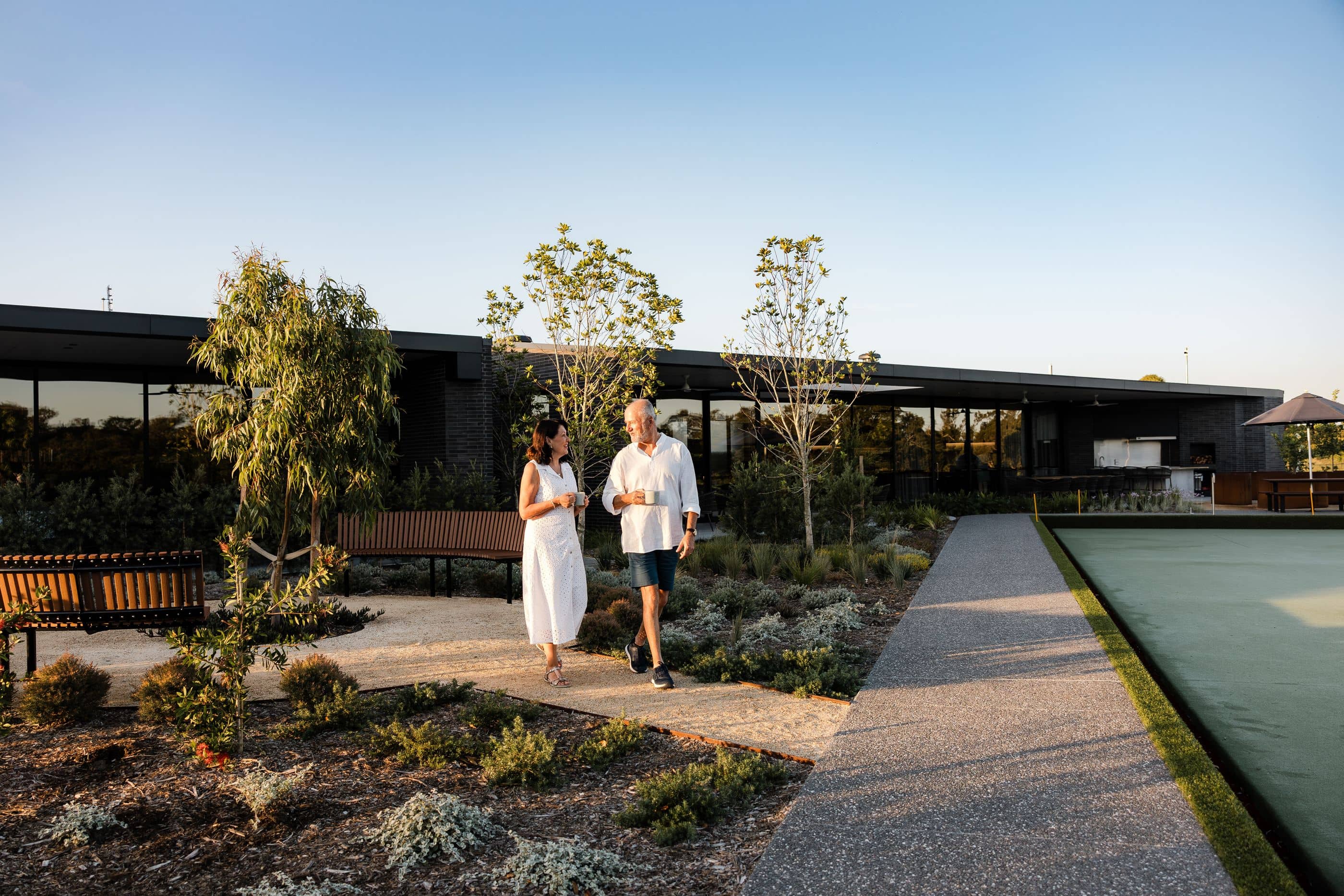 A couple walks with coffee cups in an Ingenia Lifestyle community by a bowling green.