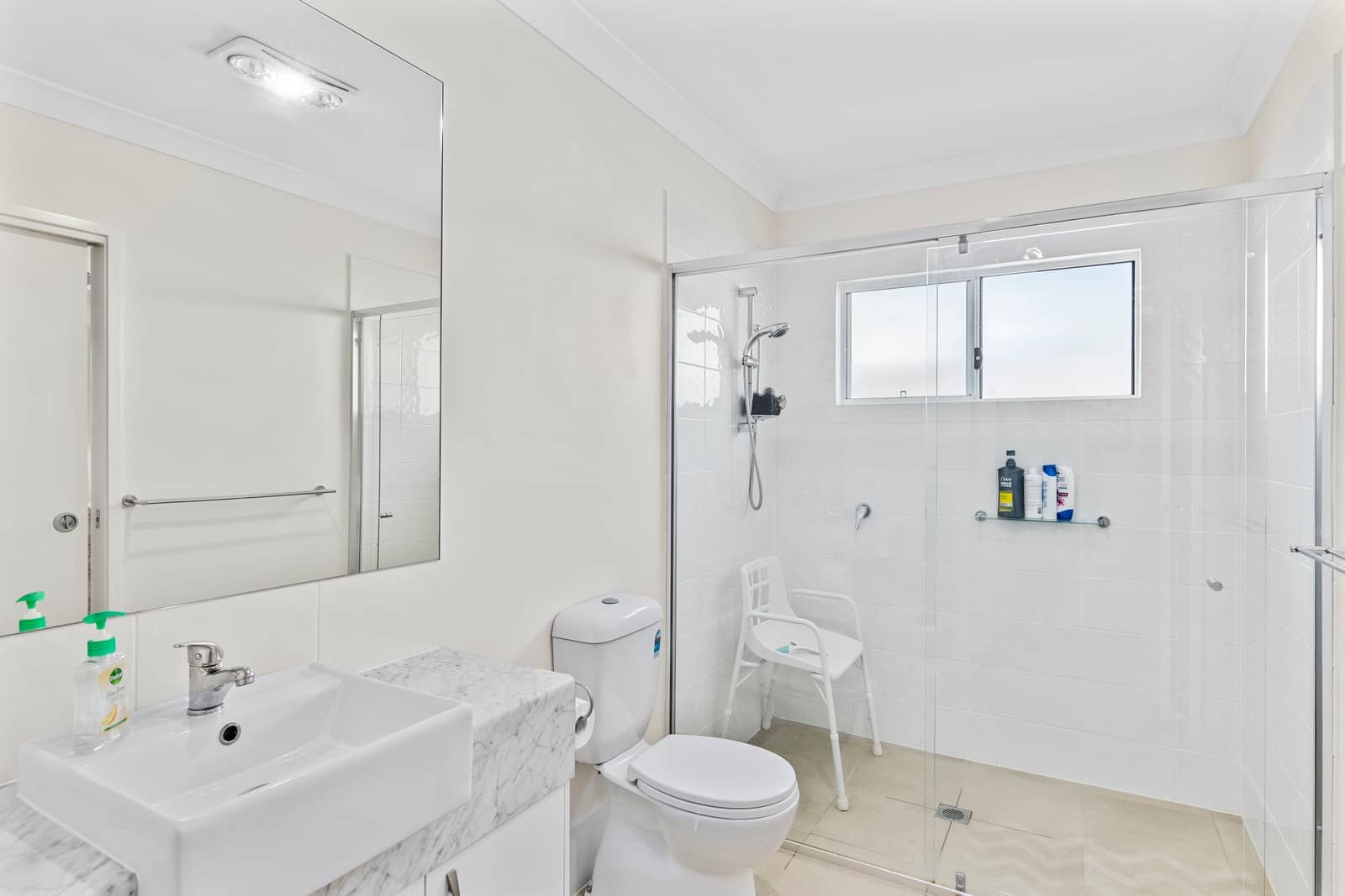 A modern, accessible bathroom in an Ingenia Lifestyle community featuring a white toilet, a large vanity with a marble-look benchtop and a white basin, and a glass-enclosed shower with a shower chair.