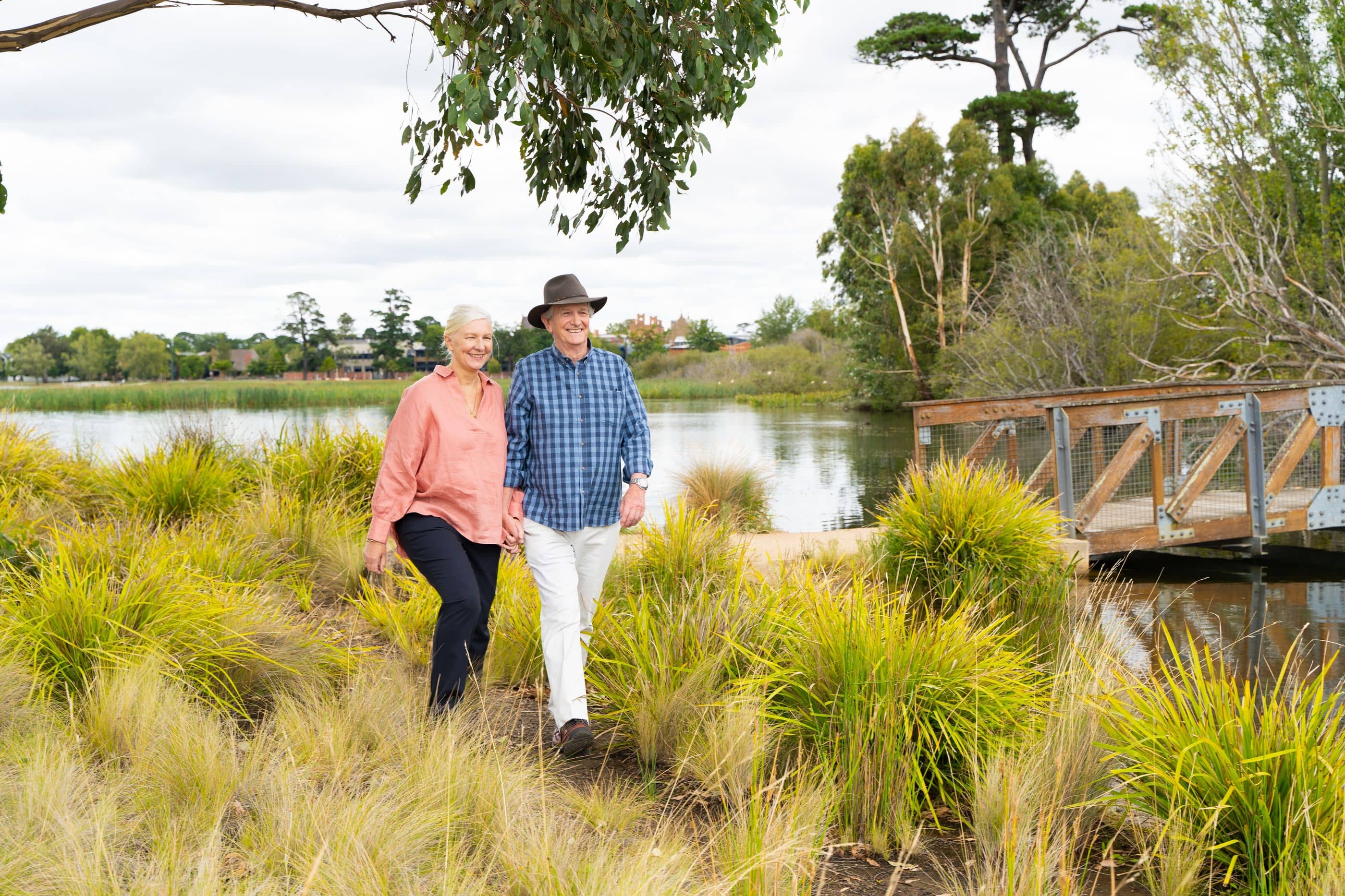 Two smiling people holding hands, walking through landscaped grounds beside a lake in an Ingenia Lifestyle community.