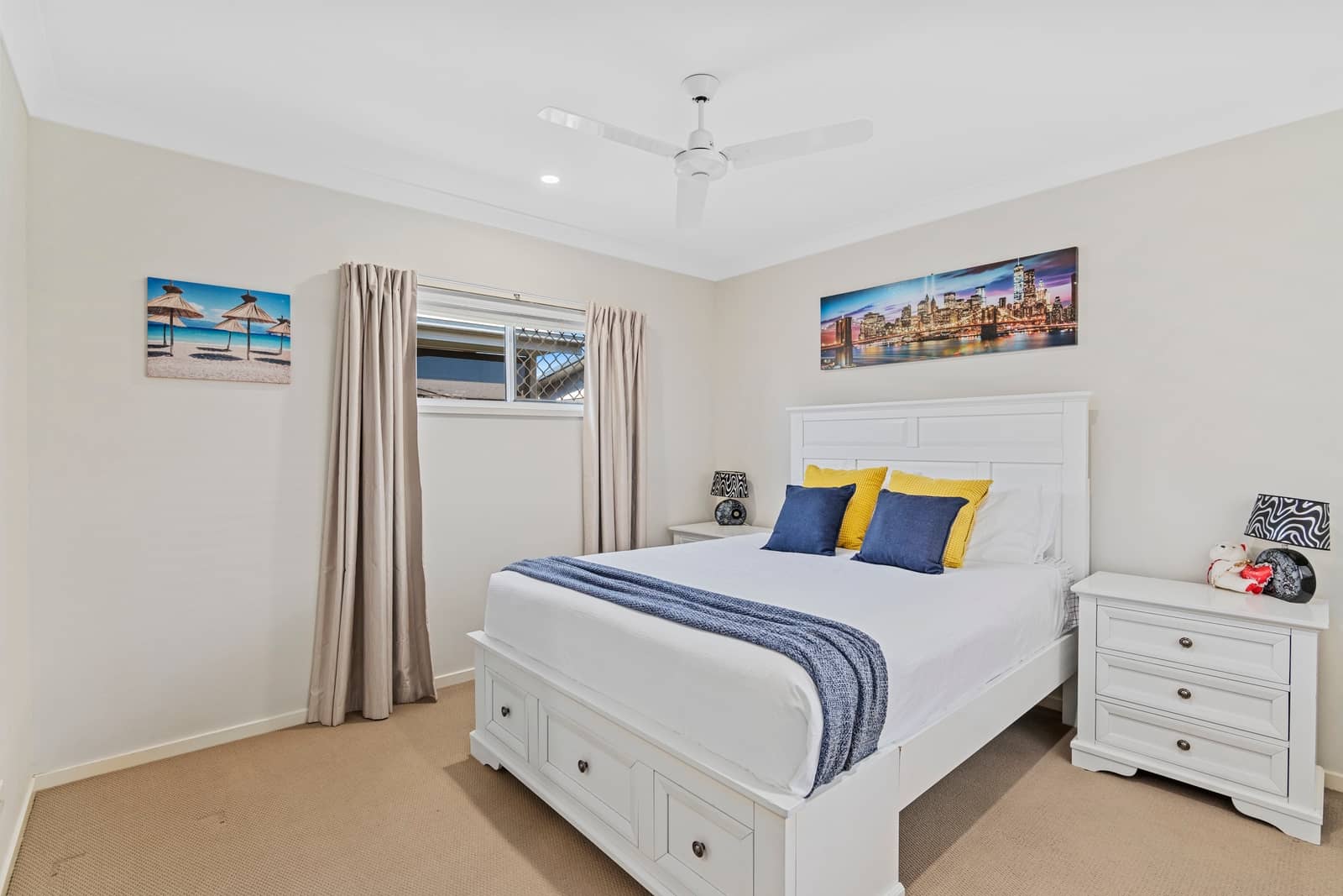 A white bed with blue and yellow pillows and a blue throw in a bedroom with two wall artworks and a ceiling fan.