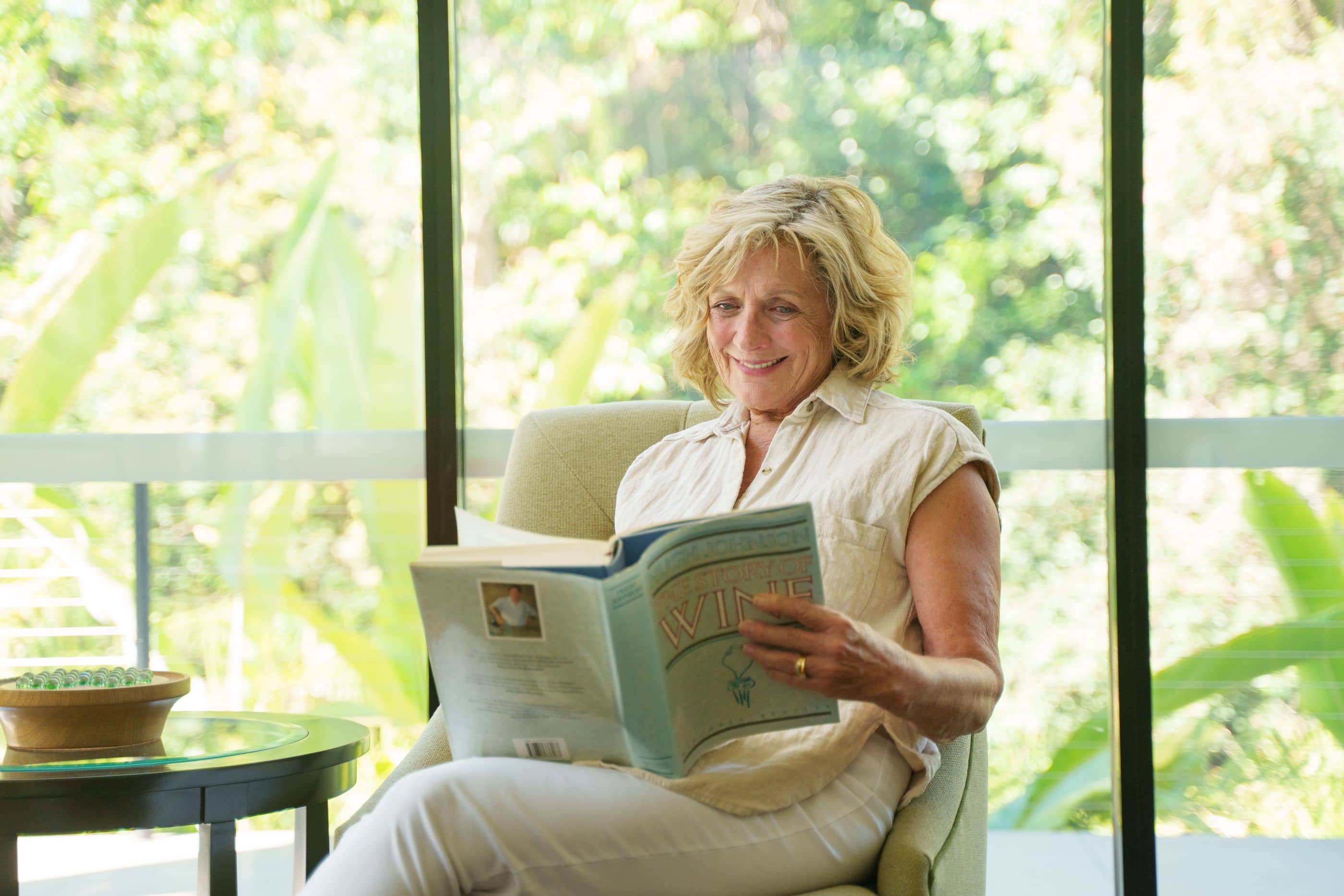 A woman smiles while reading a book in a chair next to a large window overlooking lush greenery.