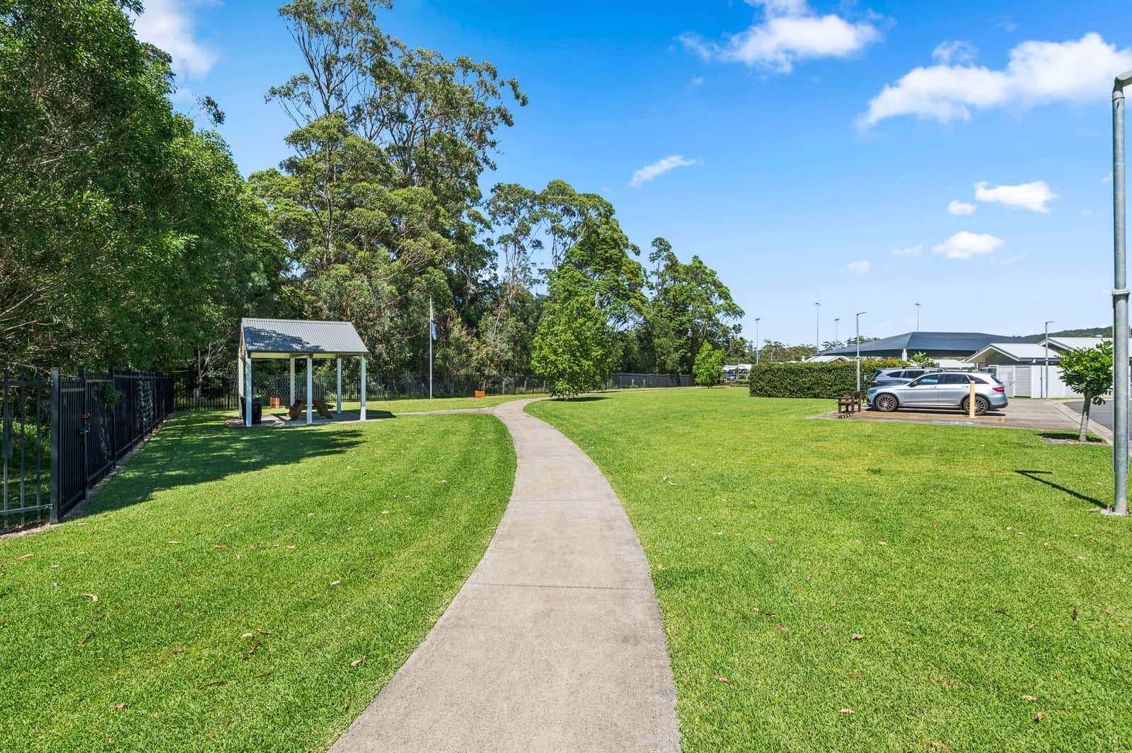 A paved path winds through a grassy common area with a covered picnic shelter on the left and homes in the background.