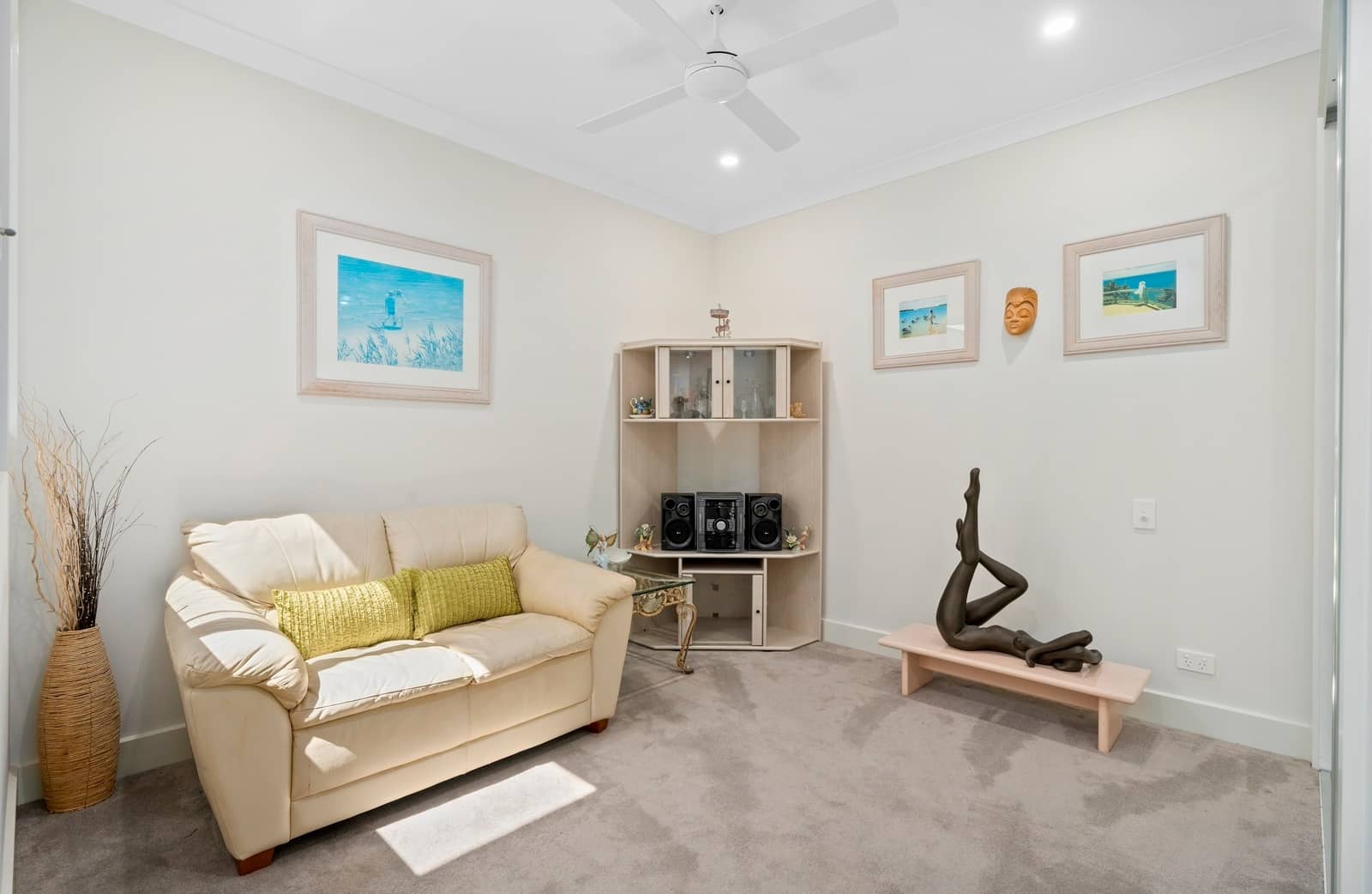 Living area with a cream sofa, light carpet, corner shelving unit, coastal artwork, and ceiling fan, in an Ingenia Lifestyle home.
