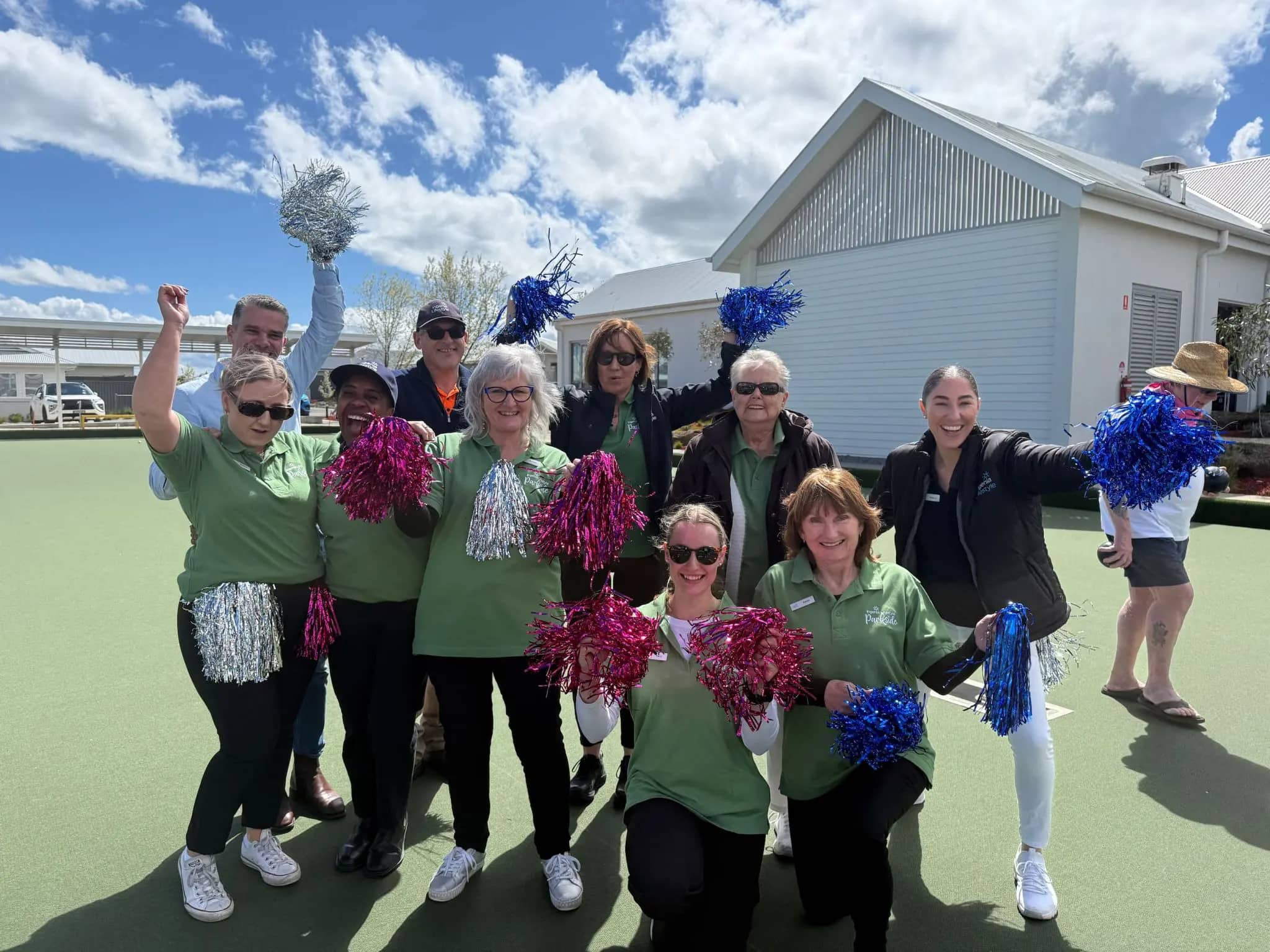 A group of people in green shirts and black pants pose with pom-poms on a green surface, with a modern building and blue sky behind them.
