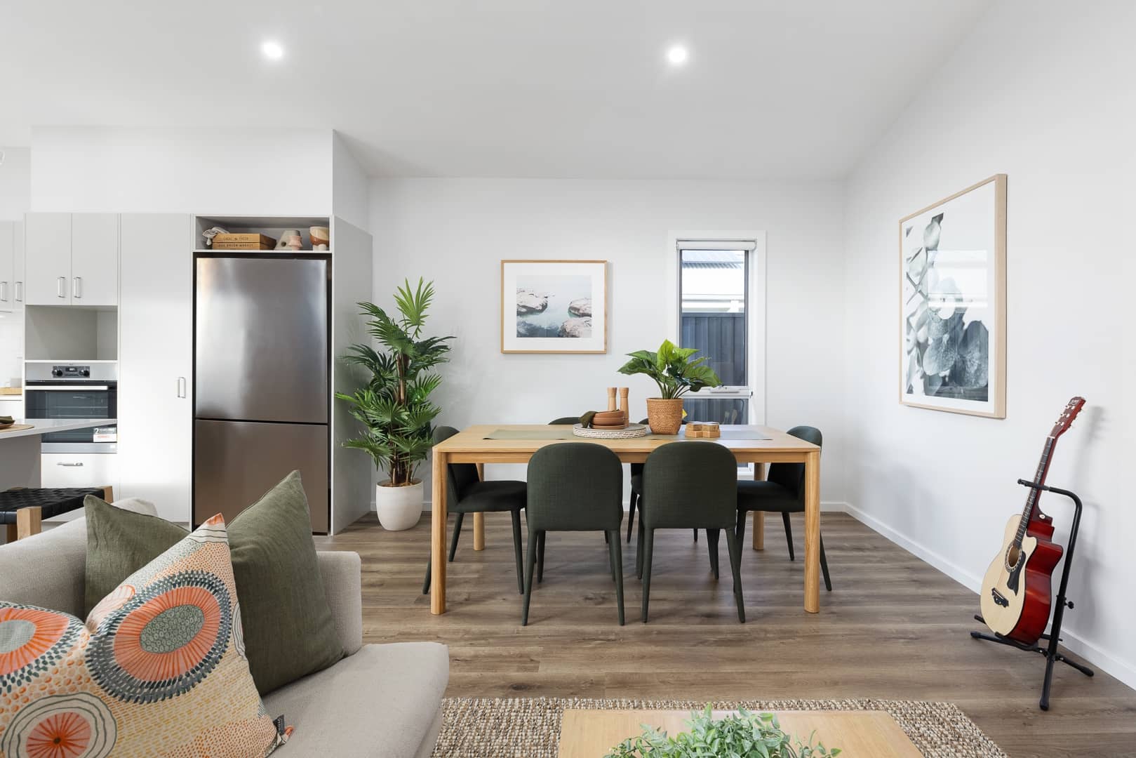 A modern dining and living area in an Ingenia Lifestyle home, featuring a wooden dining table with chairs, a sofa, a refrigerator, and artwork.