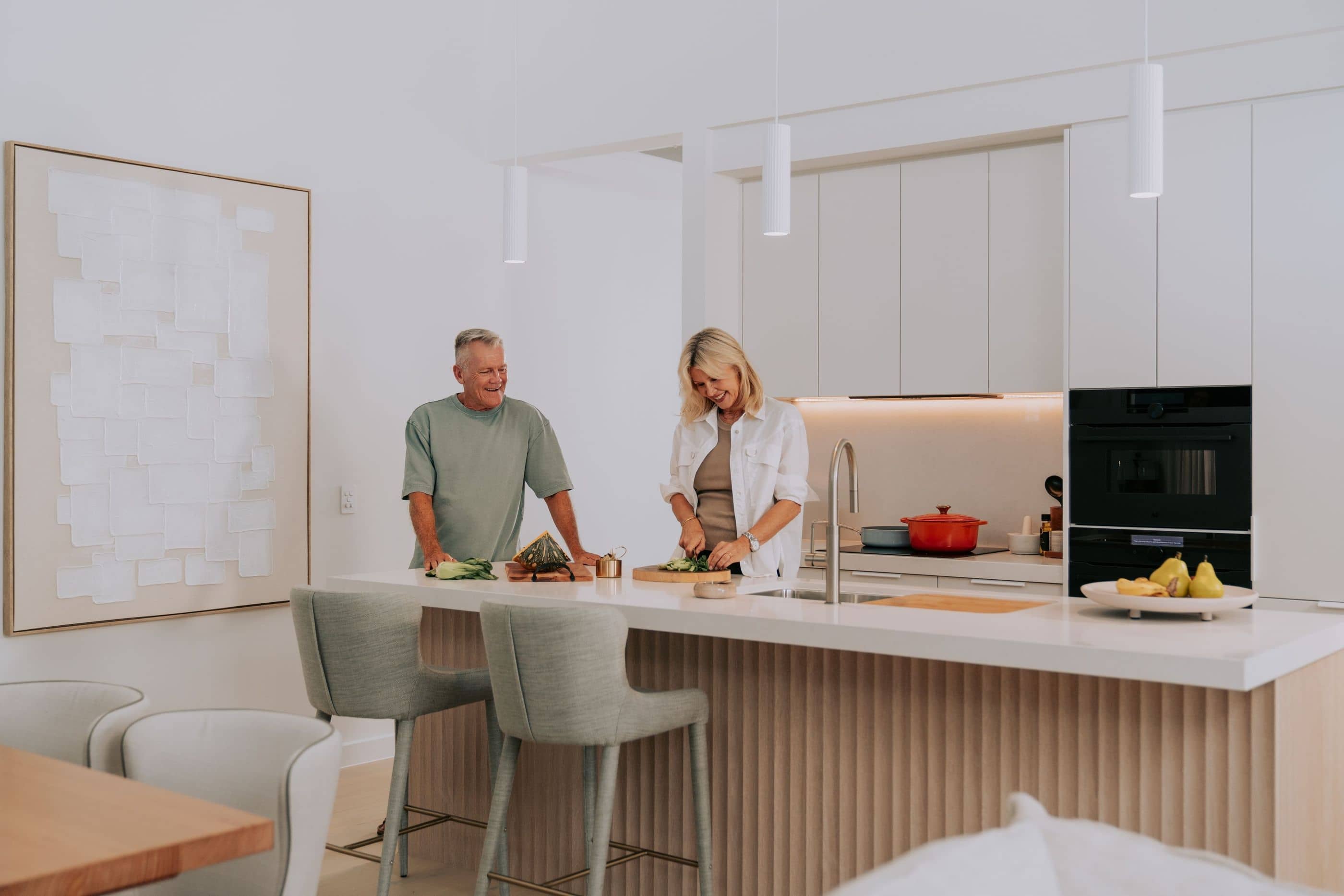 A man and woman smiling while preparing food in a modern Ingenia Lifestyle kitchen with an island bench and contemporary appliances.