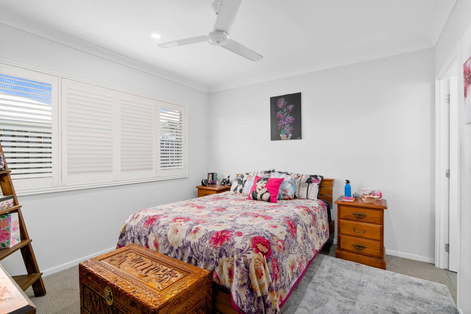 A bedroom with a floral bedspread, decorative pillows, white plantation shutters, and a wooden chest at the foot of the bed.