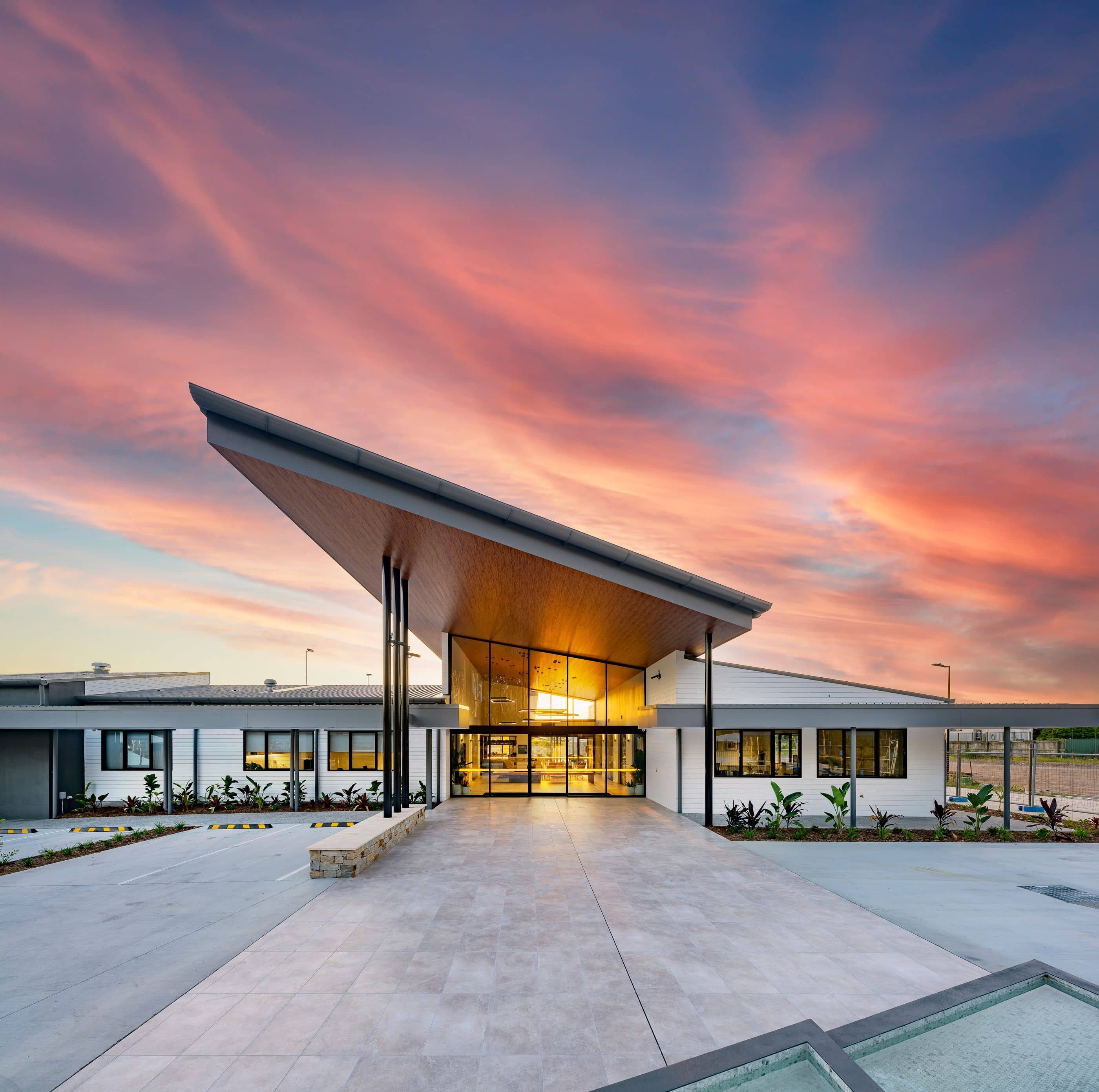 Modern Ingenia Lifestyle community clubhouse with an illuminated glass entrance, angular roof, and landscaped grounds at sunset.