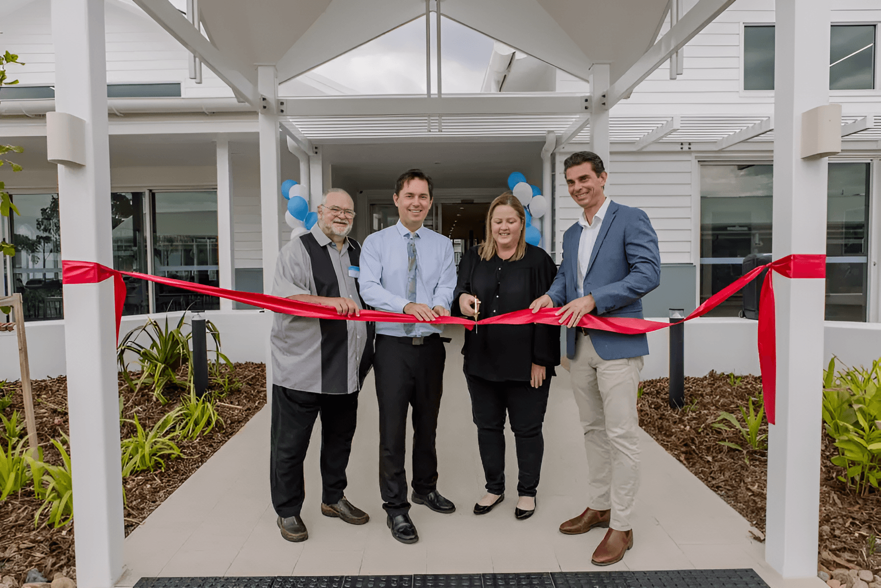 Four people participate in a ribbon-cutting ceremony in front of a white building with blue and white balloons.