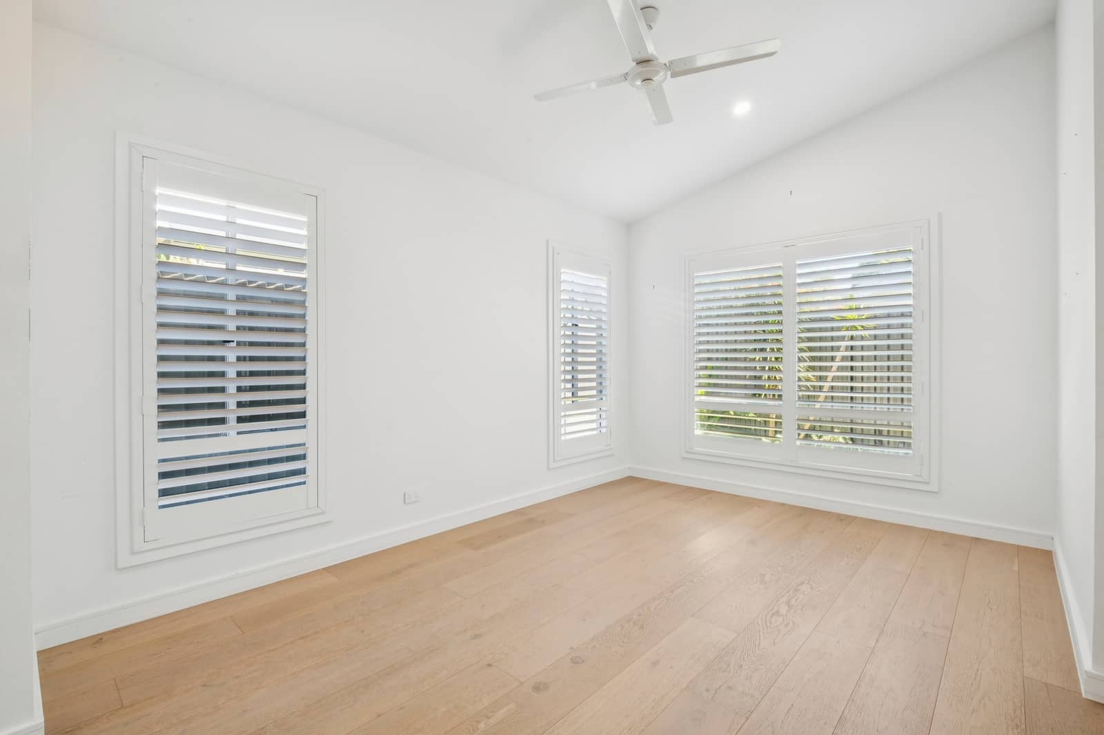 Interior view of an empty, bright room with white walls, a vaulted ceiling, and a ceiling fan. Two windows with white plantation shutters look out onto greenery. The floor is light wood.