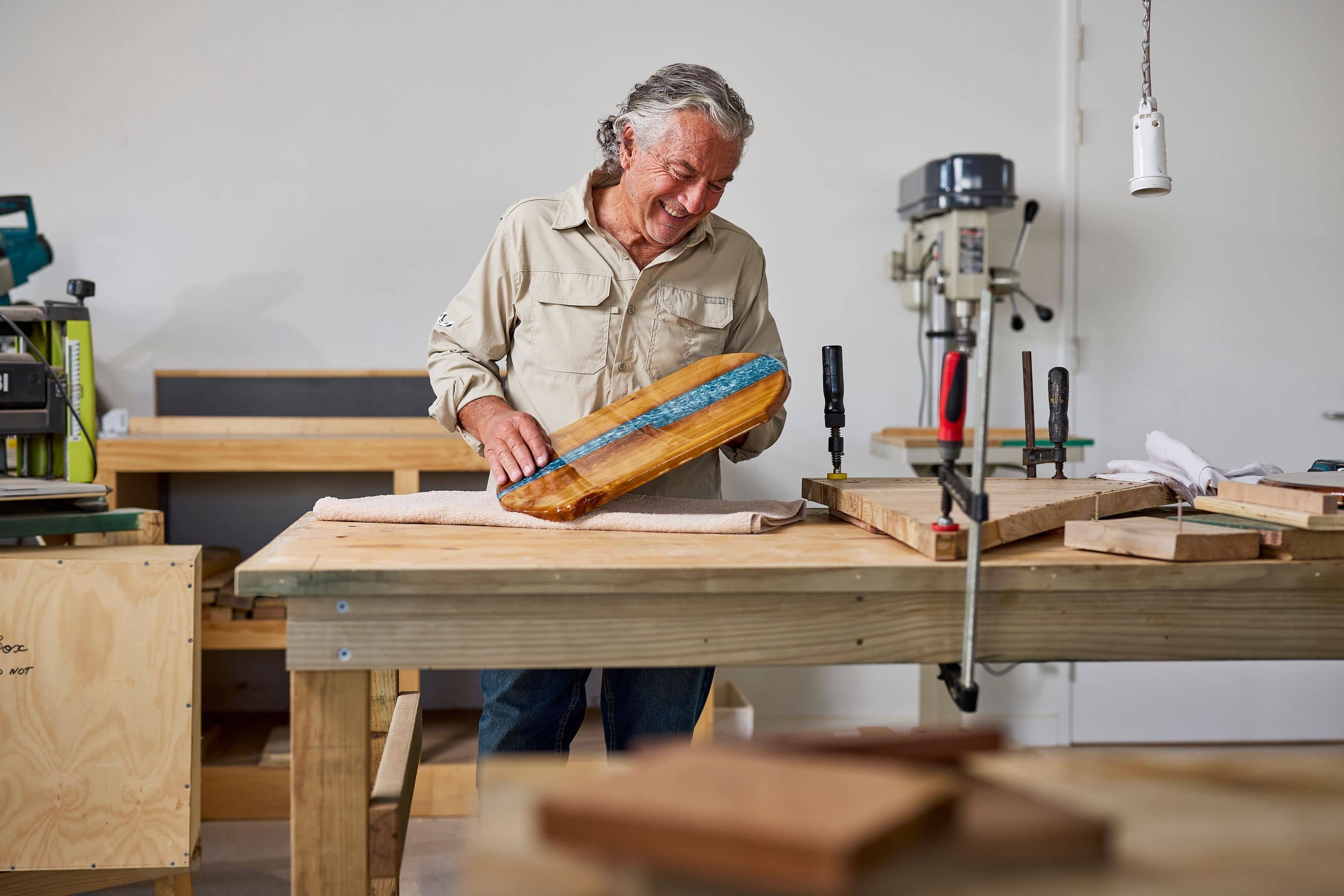 Man in tan shirt smiling and holding a wooden board with blue resin inlay in a workshop.