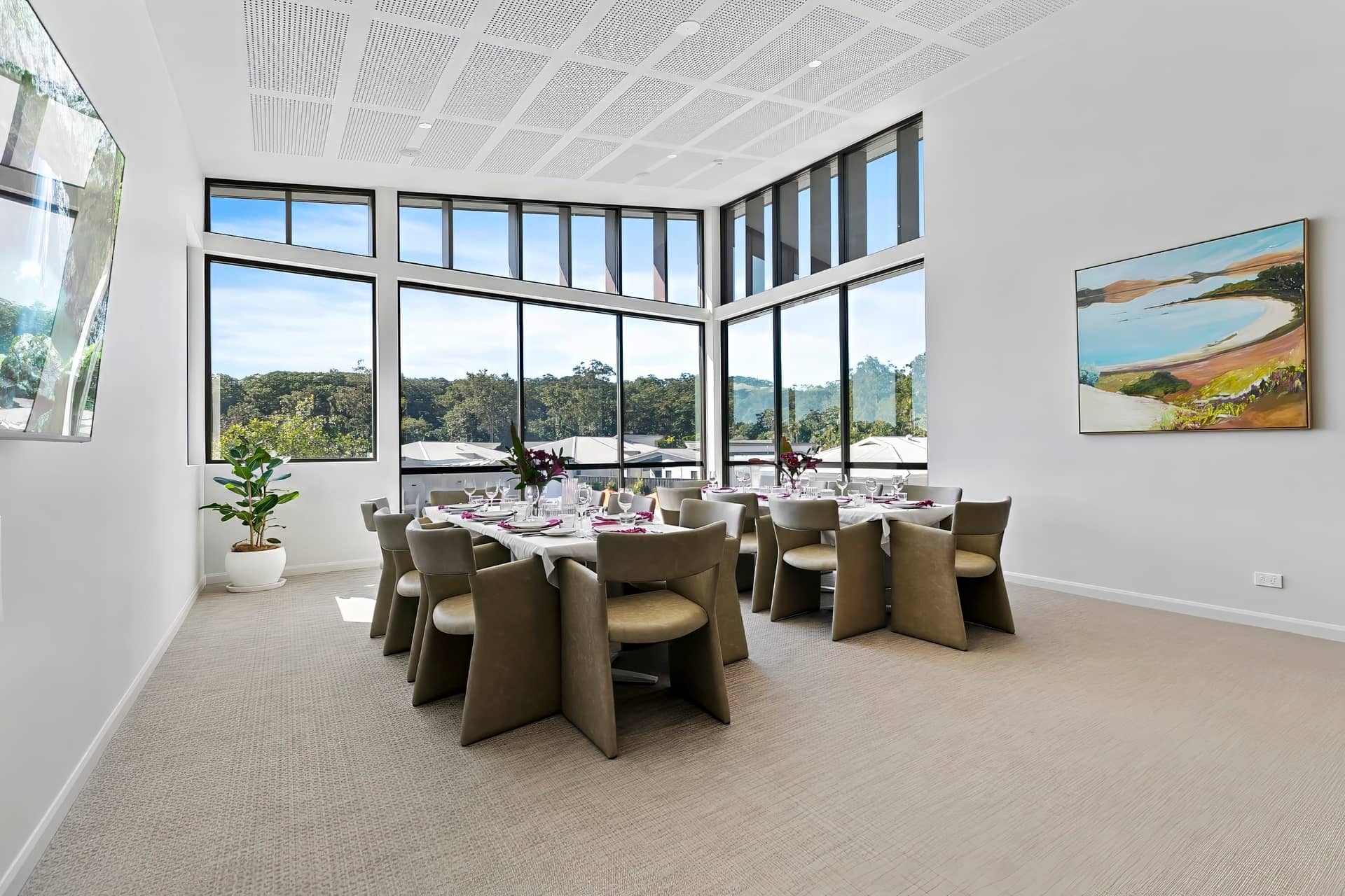 Ingenia Lifestyle community dining area with two tables set, large windows viewing trees, and an abstract painting.