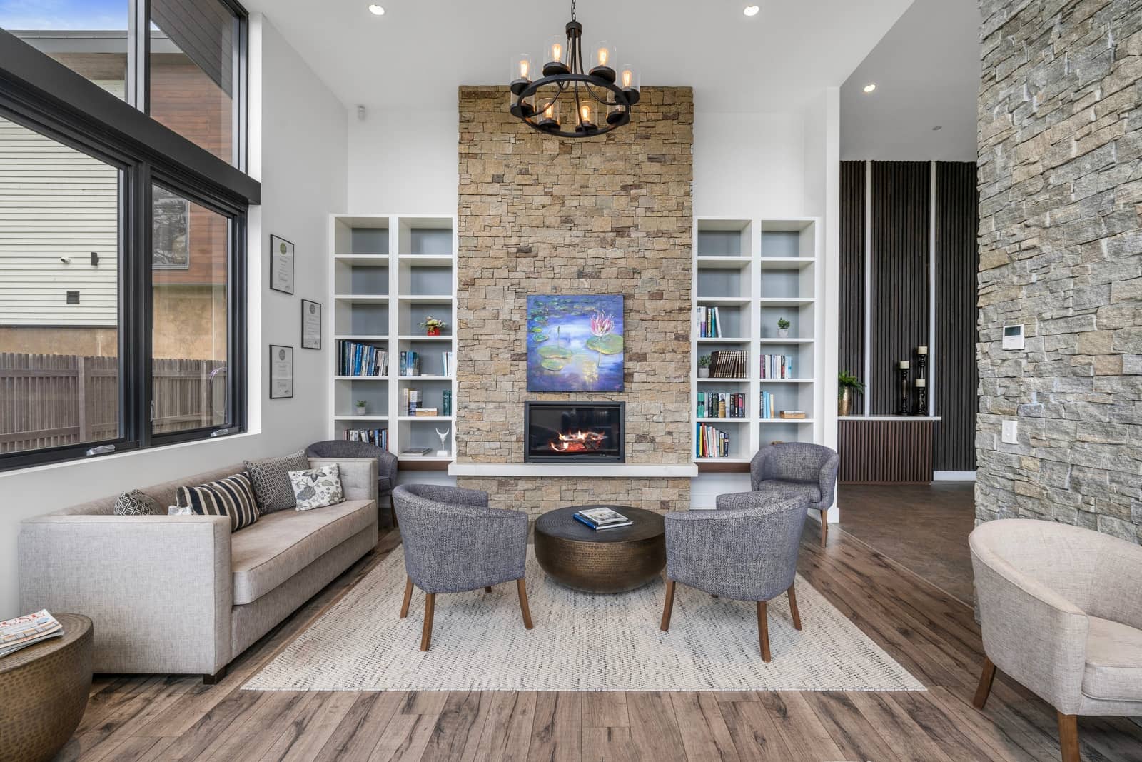 An Ingenia Lifestyle community lounge featuring a stone fireplace, built-in bookshelves, various seating, and tall windows.