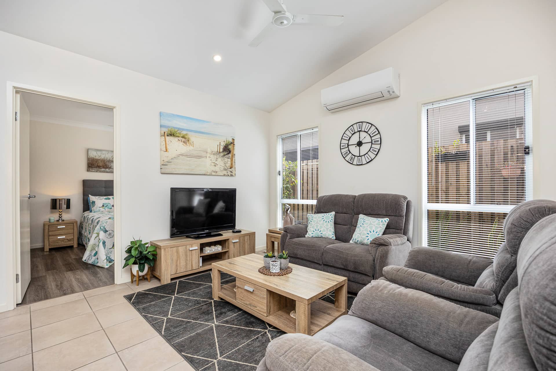 Bright living room in an Ingenia Lifestyle home with gray recliners, wooden coffee table, TV, and a view into a bedroom.