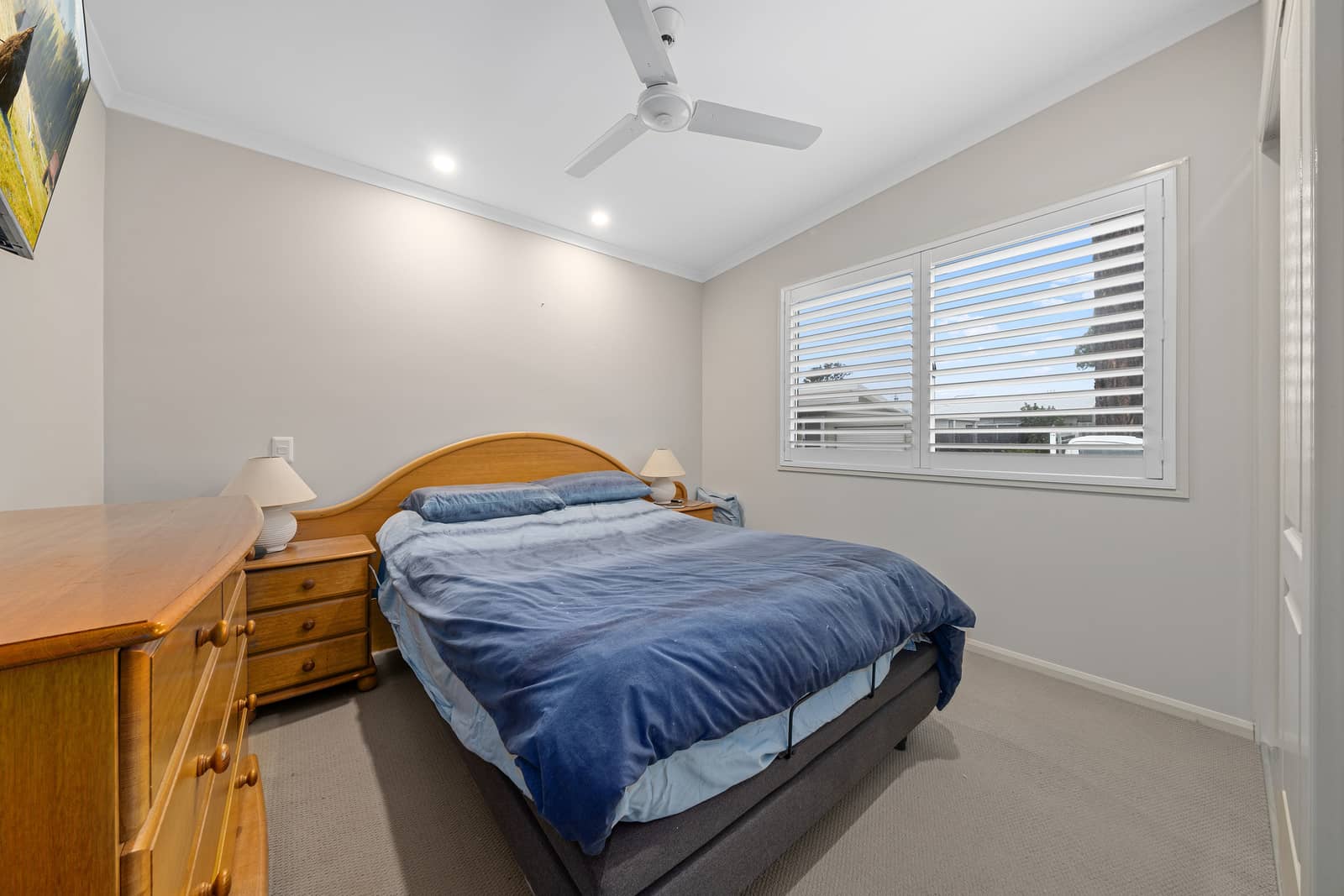Bedroom in an Ingenia Lifestyle home with bed, wooden furniture, window with white shutters, and ceiling fan.