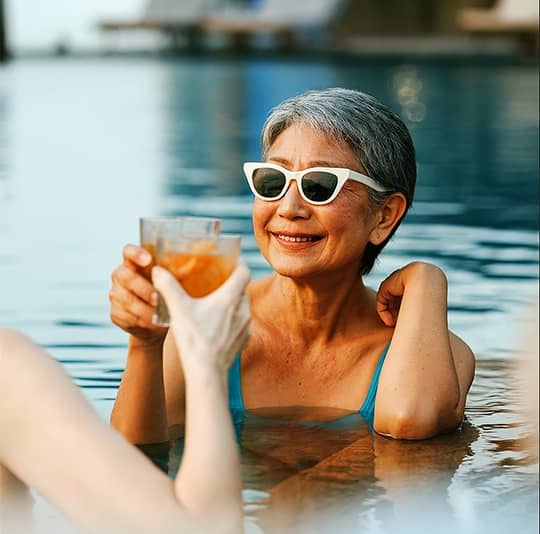 Smiling woman in white sunglasses in a swimming pool, clinking drinks, enjoying community amenities.